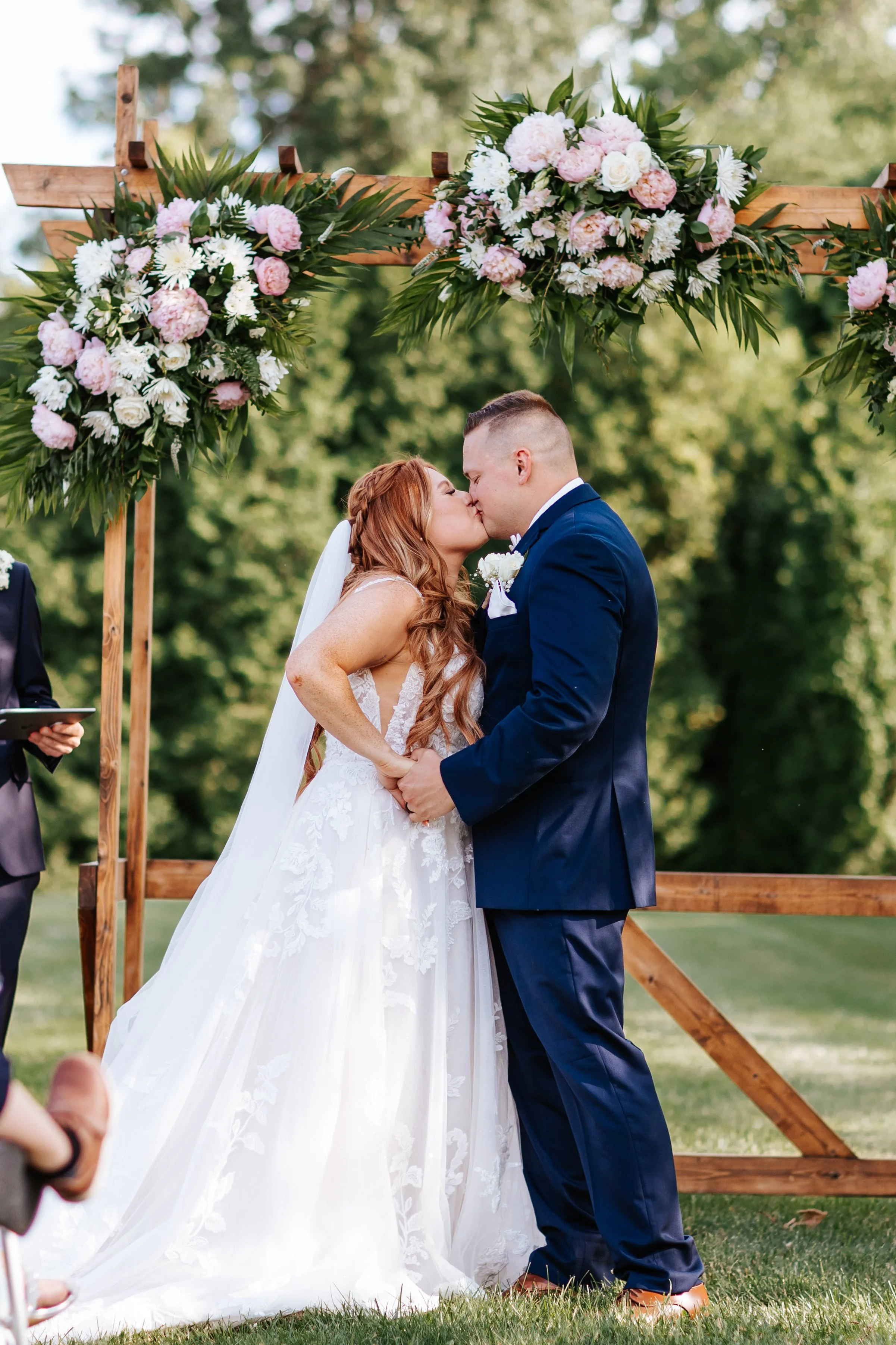 Bride and groom kissing during their outdoor wedding ceremony under a wooden arch decorated with pink and white flowers and green foliage.