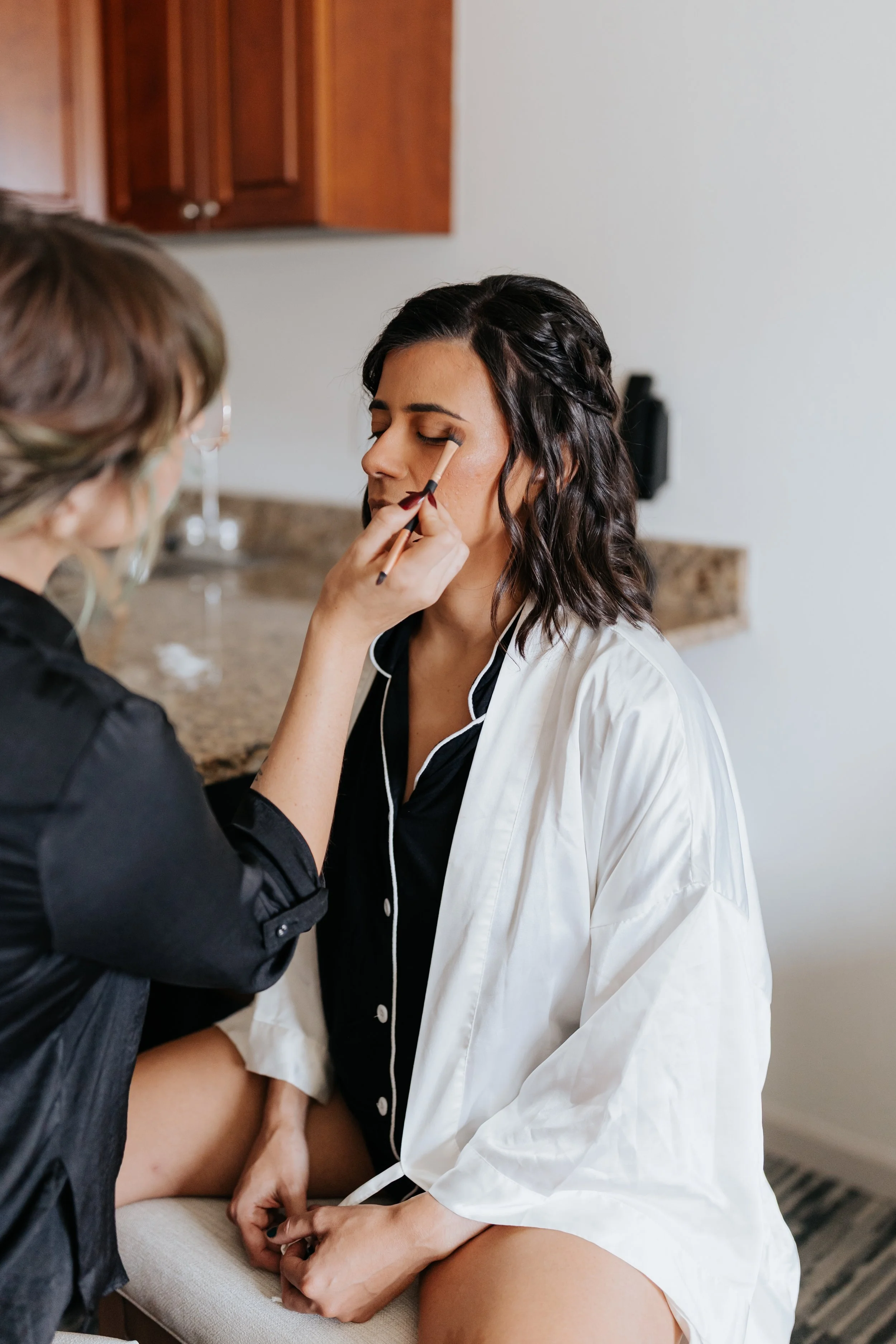A woman with dark wavy hair getting her makeup done by a makeup artist in a black robe, sitting with eyes closed in a bedroom or dressing area.