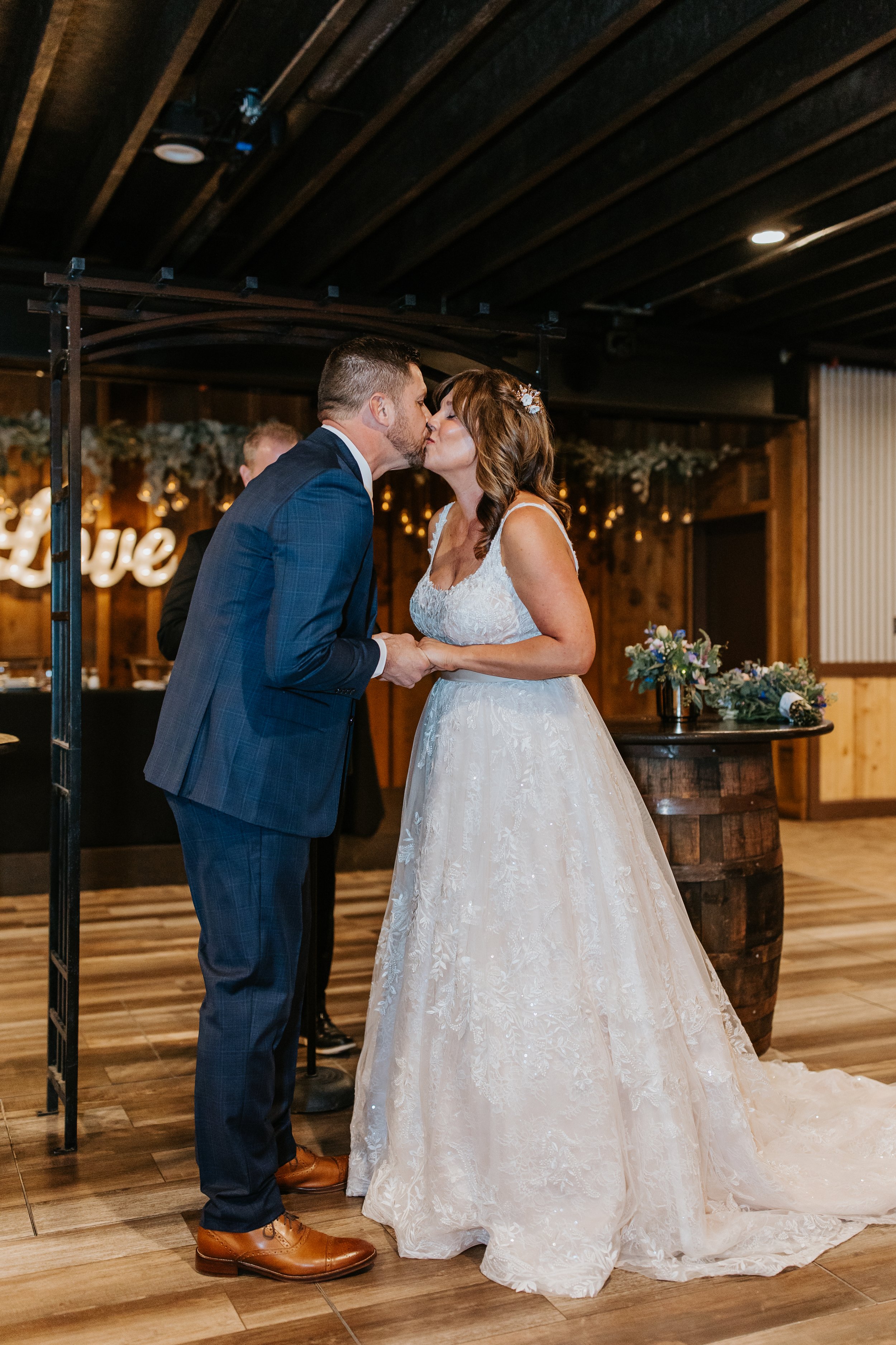 A bride and groom share a kiss during their wedding reception in a rustic venue with wood accents and floral decorations.