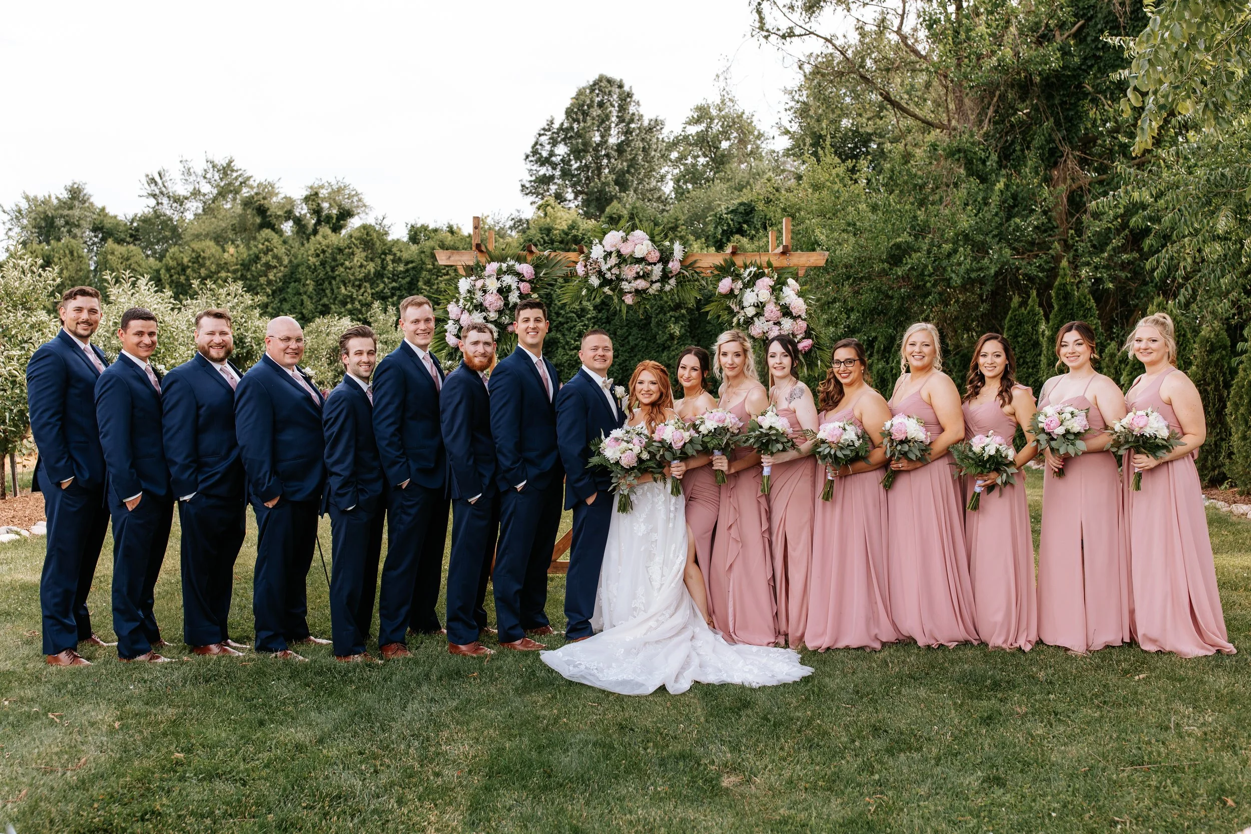 A wedding party standing outdoors on grass in front of a floral arch, with 9 men in navy suits and 7 women in pink dresses, holding bouquets of flowers.