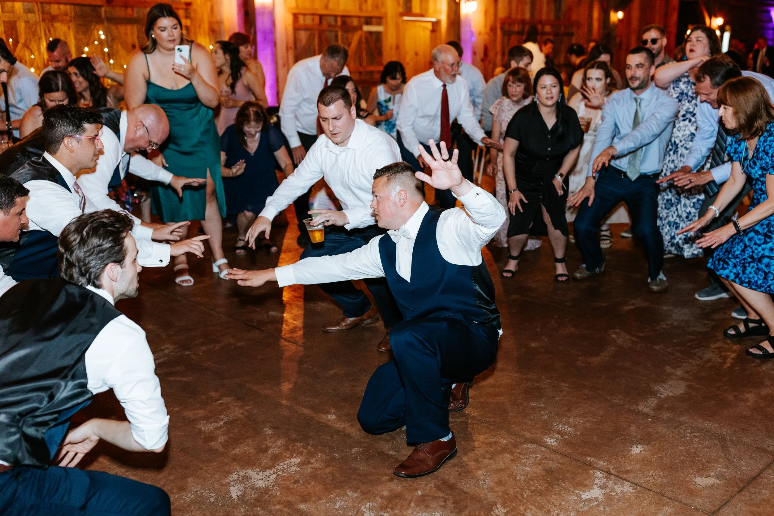 People dancing at a party or wedding reception, with some forming a circle and others dancing in the center, on a wooden dance floor with colorful lighting.