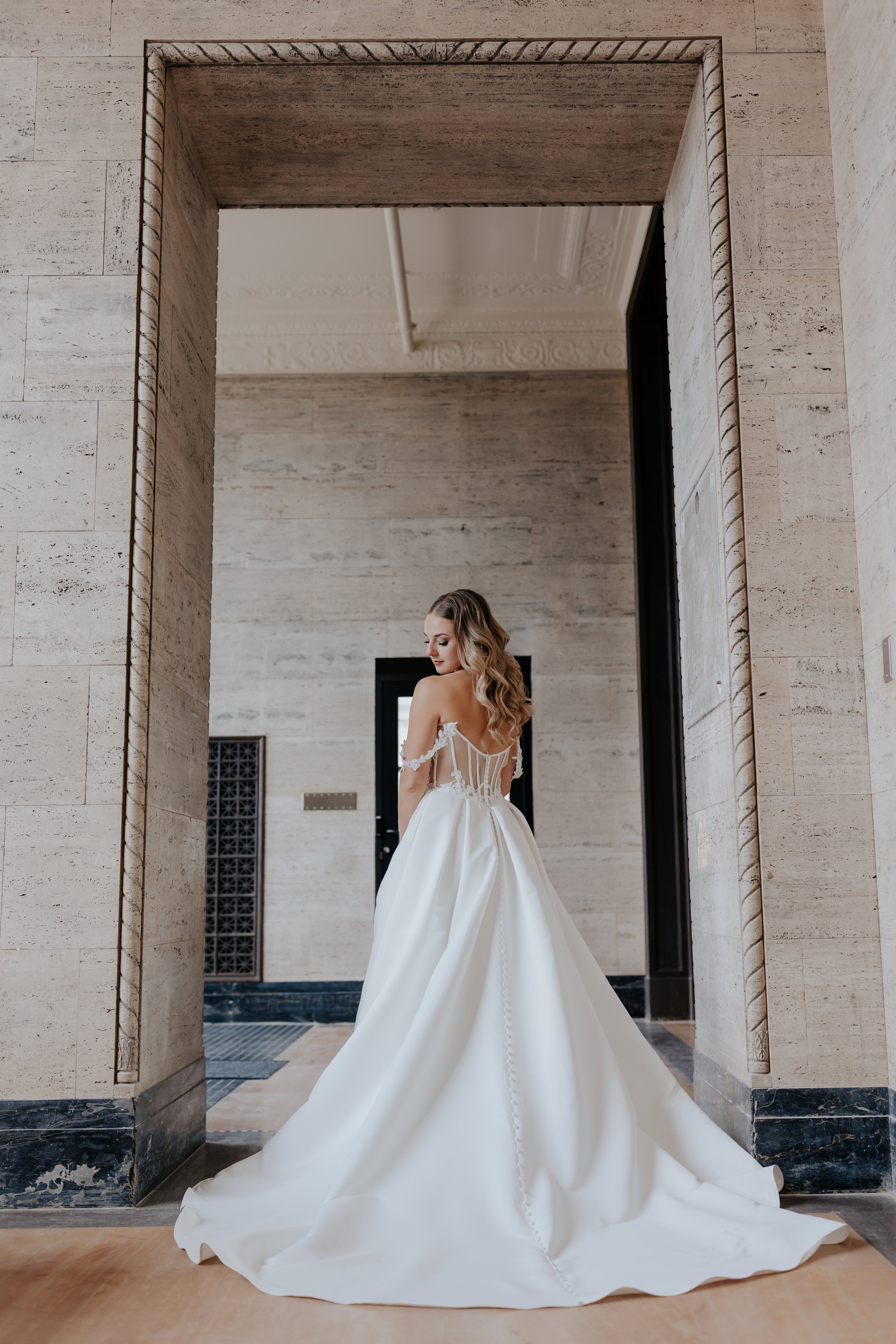 A bride in a wedding gown standing in an elegant, modern interior through a stone archway.