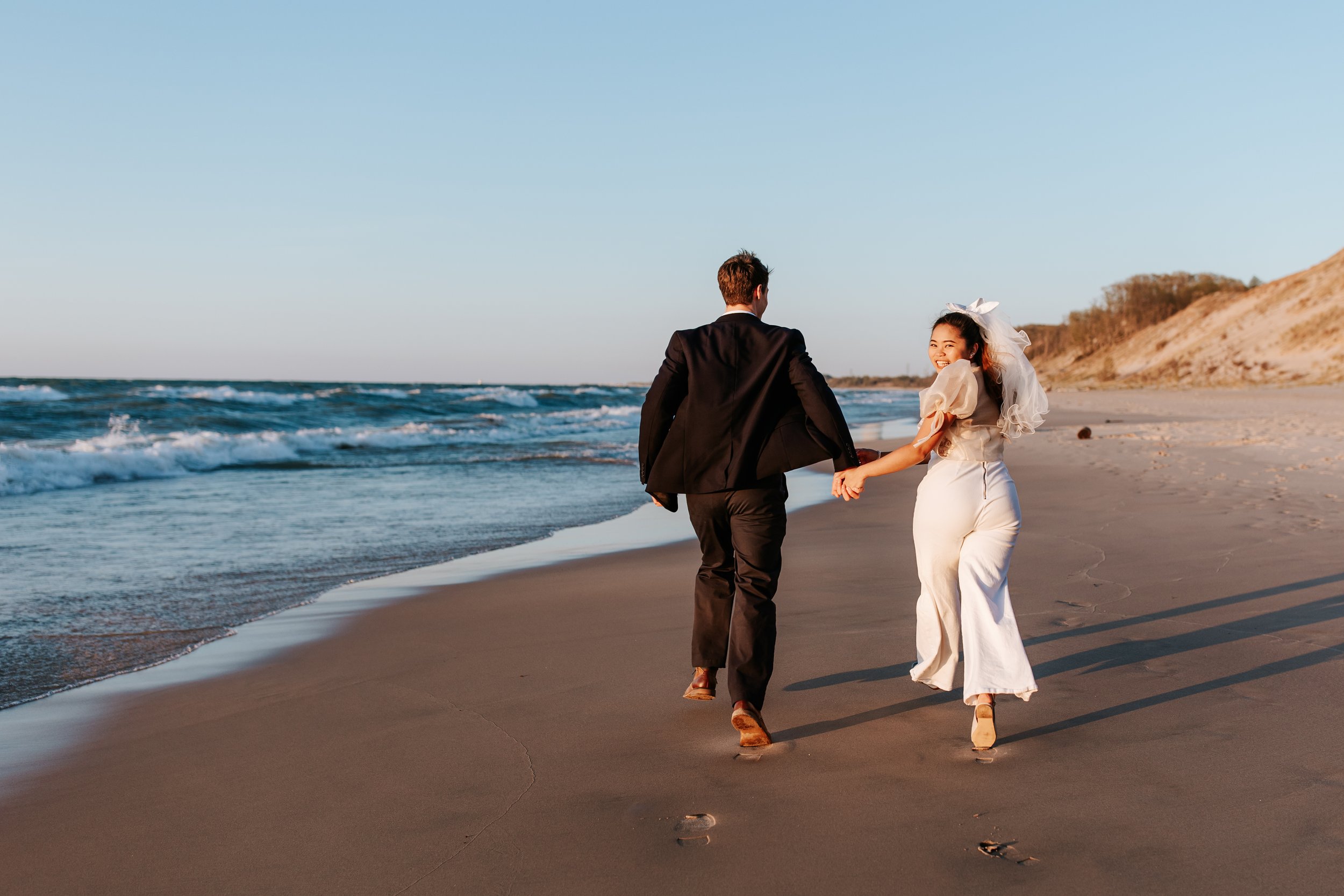 A couple in wedding attire holding hands and walking on a beach, with the ocean waves and dunes in the background.