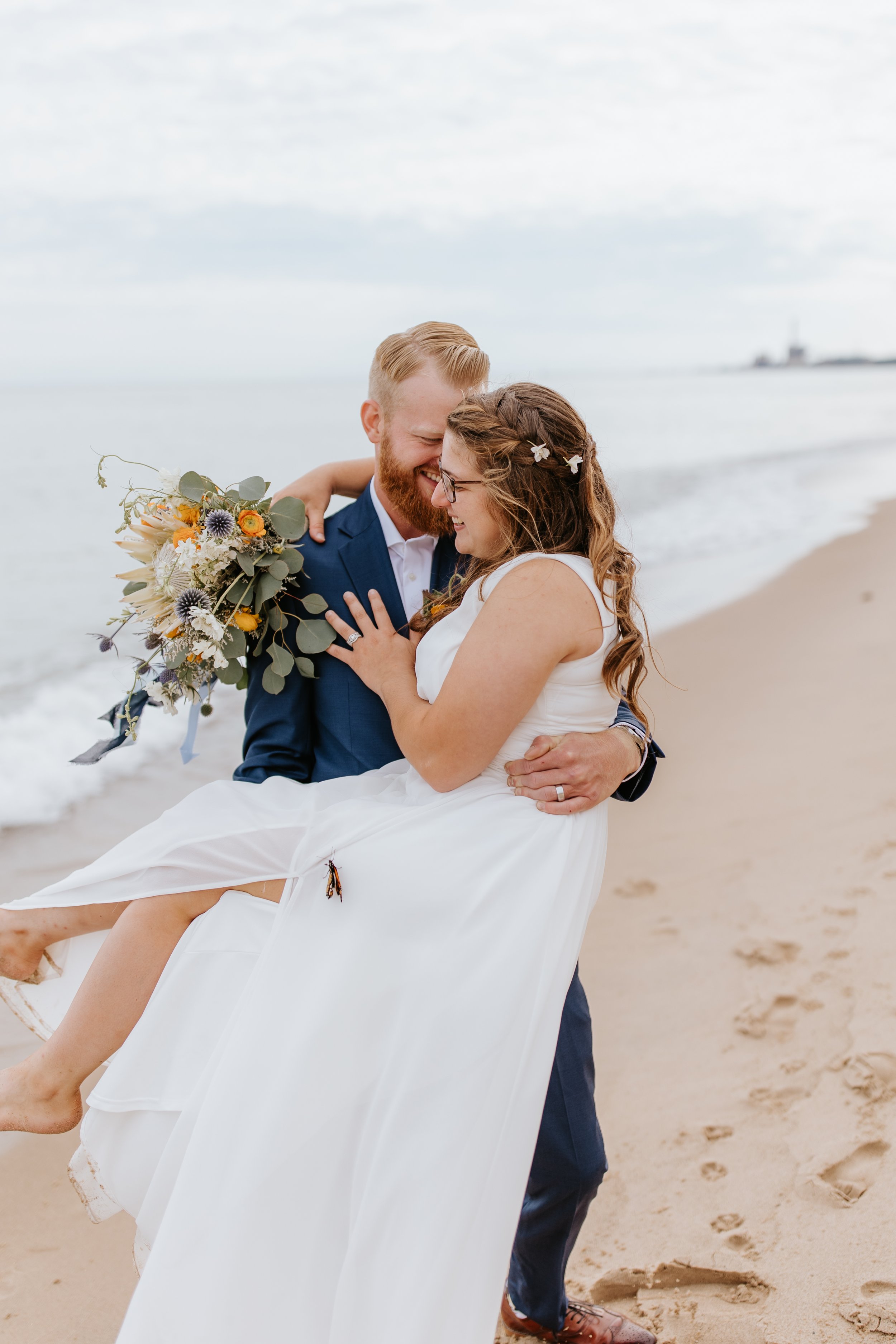 A bride being carried by a groom on the beach, both smiling and about to kiss, with a bouquet in her hand.
