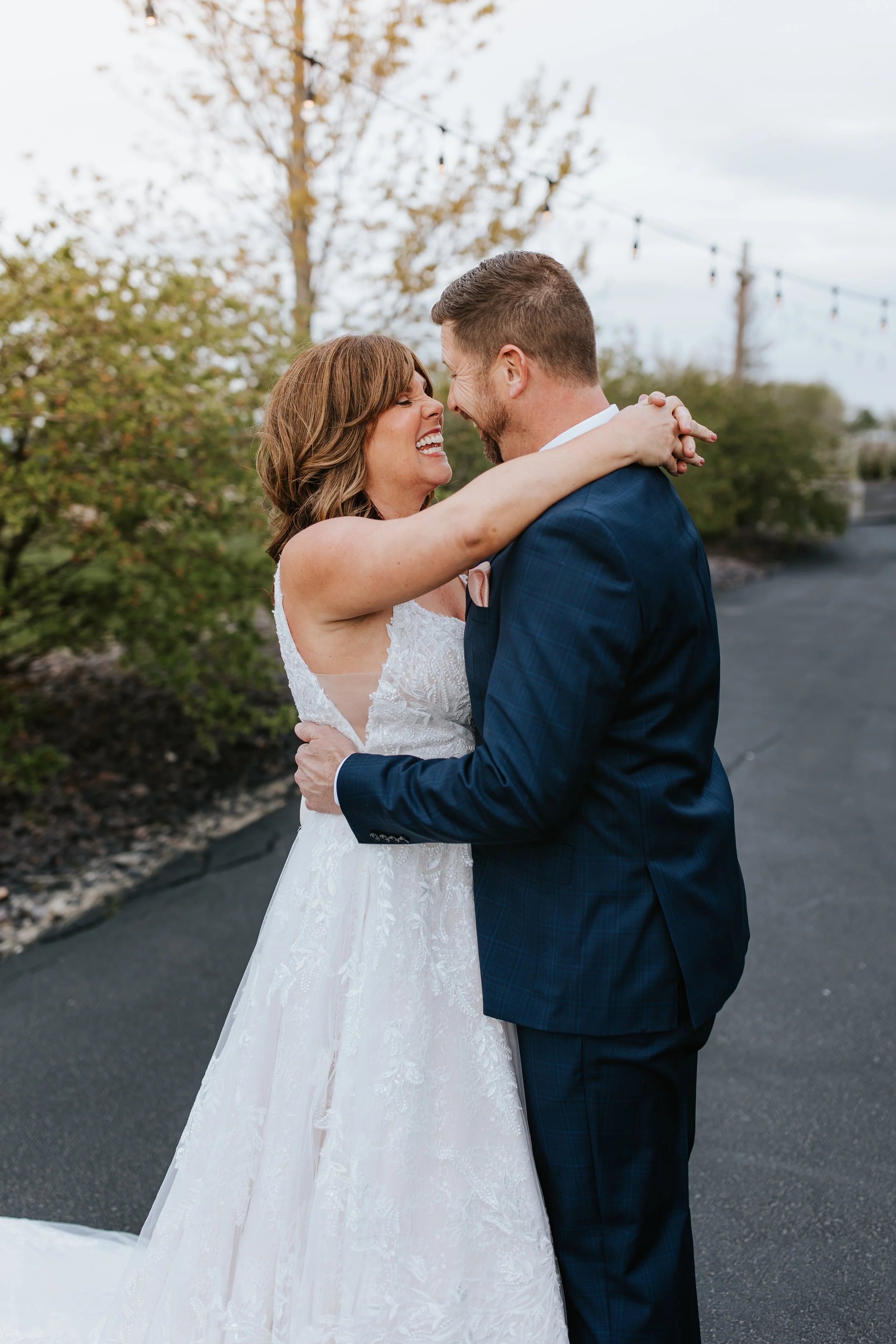 A bride and groom embrace and smile outdoors, wearing wedding attire, with trees and string lights in the background.