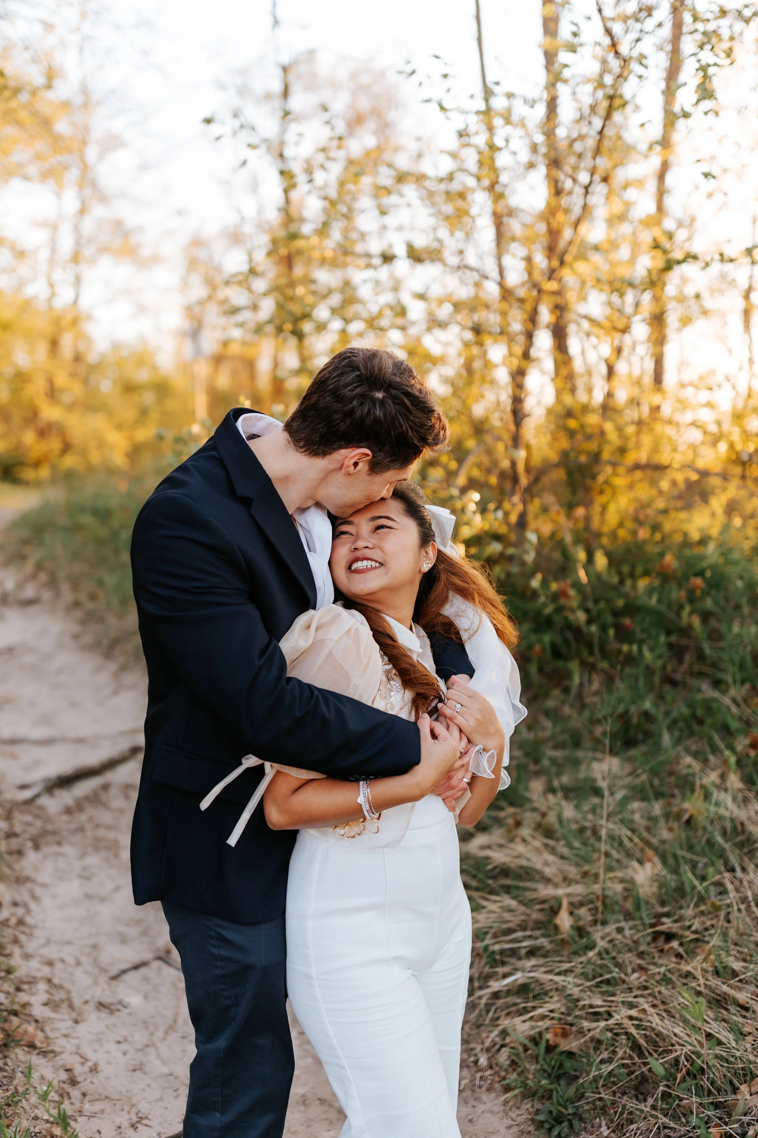 A couple hugging on a dirt path surrounded by trees with fall foliage at sunset, smiling and looking into each other's eyes.