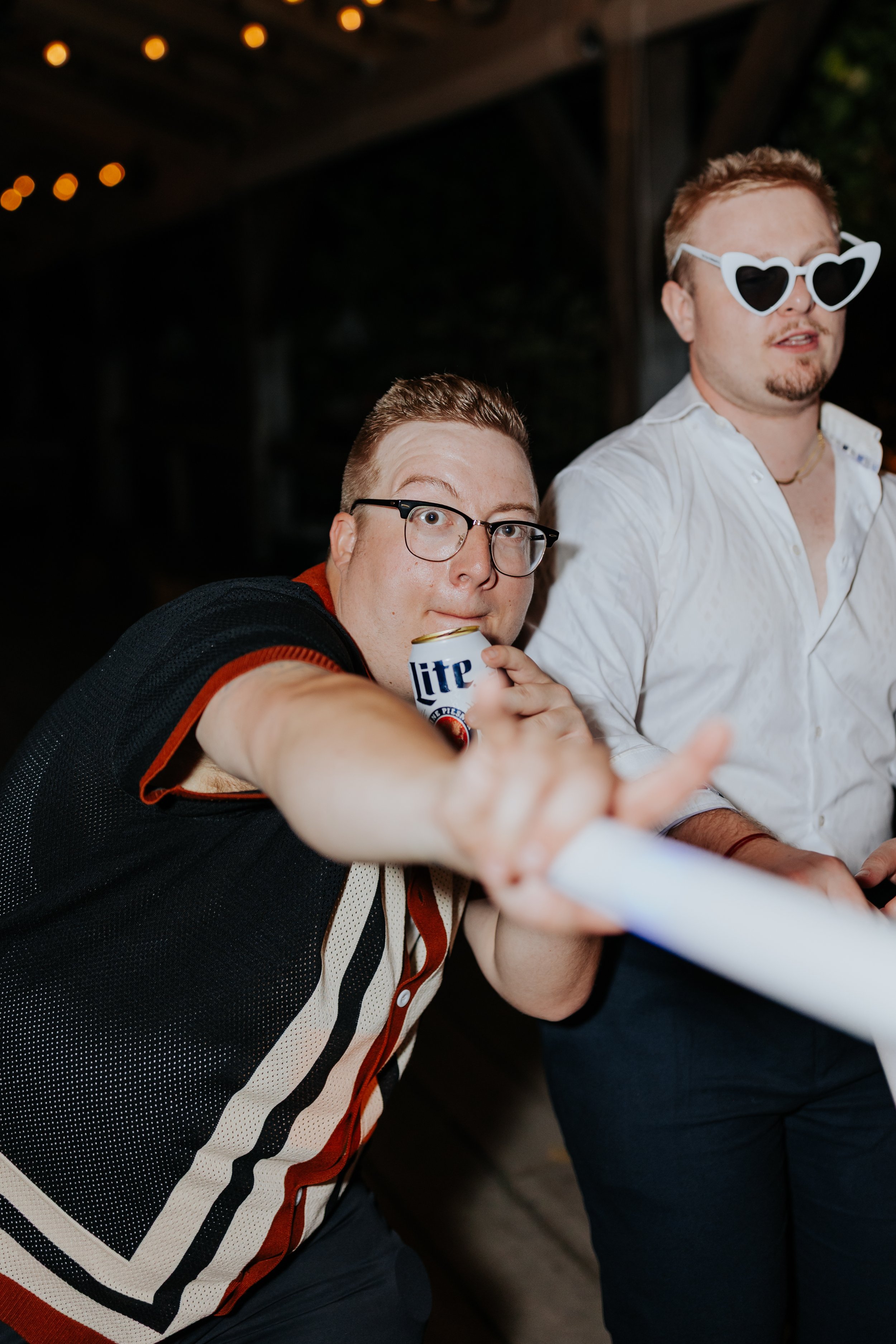 Two men at a party, one in glasses holding a Miller Lite beer can and pointing at the camera, the other in white shirt and heart-shaped sunglasses.