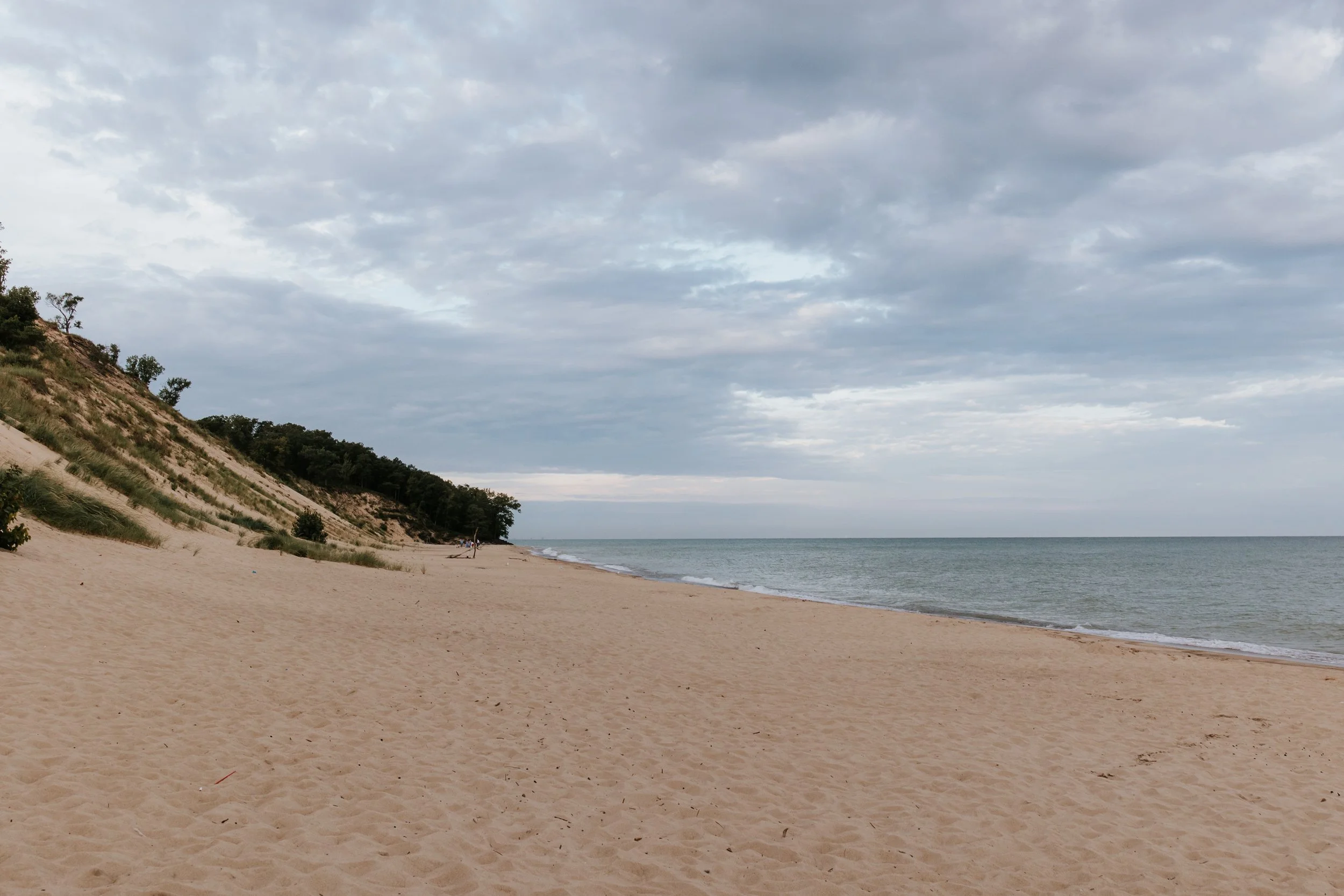 Empty sandy beach with a few small bushes and trees on a hill on the left, calm ocean water on the right, and cloudy sky overhead.