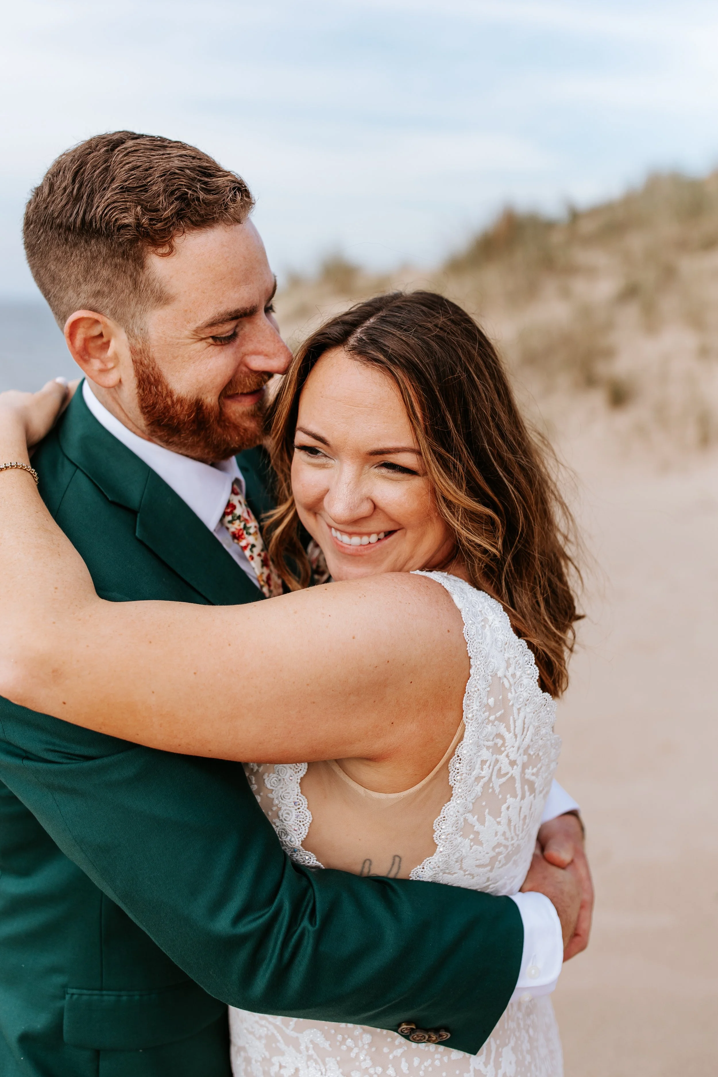A smiling couple hugging on a beach, with the woman wearing a white lace dress and the man wearing a green suit.