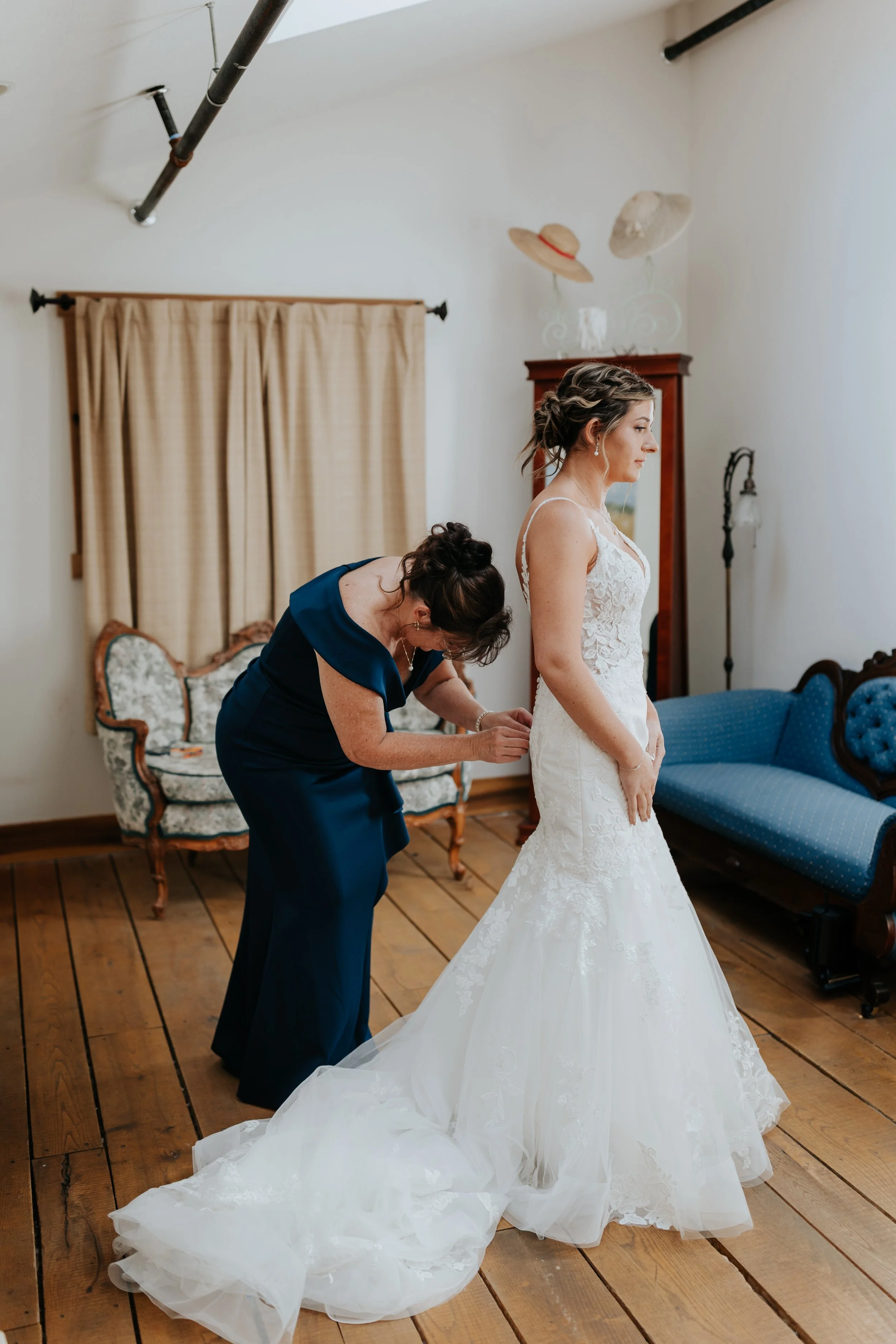 A bride in a white wedding dress having her shoes put on by a woman in a blue dress, in a room with wooden floors and vintage furniture.