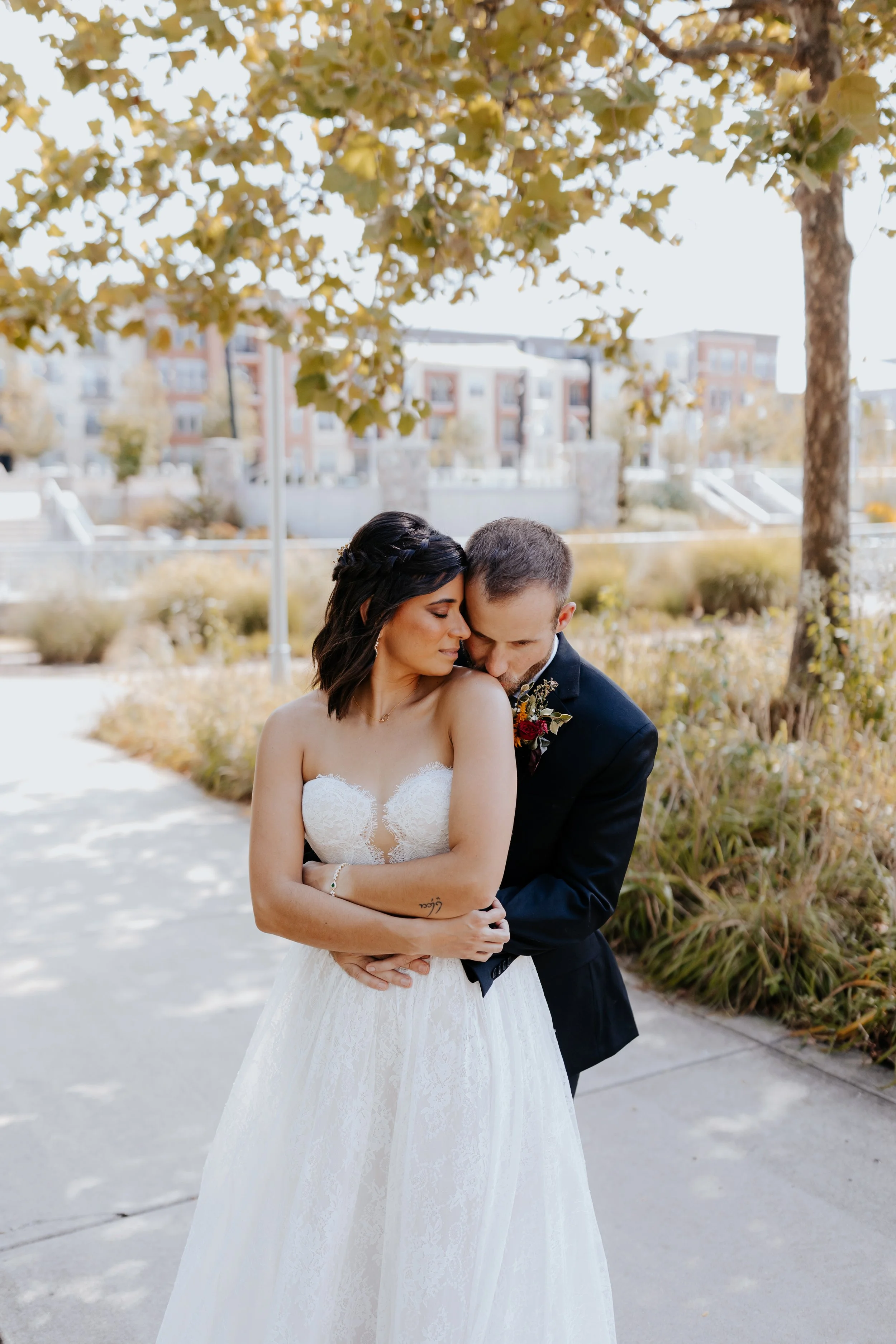 A bride and groom embrace outdoors under a tree with autumn leaves, with city buildings in the background.