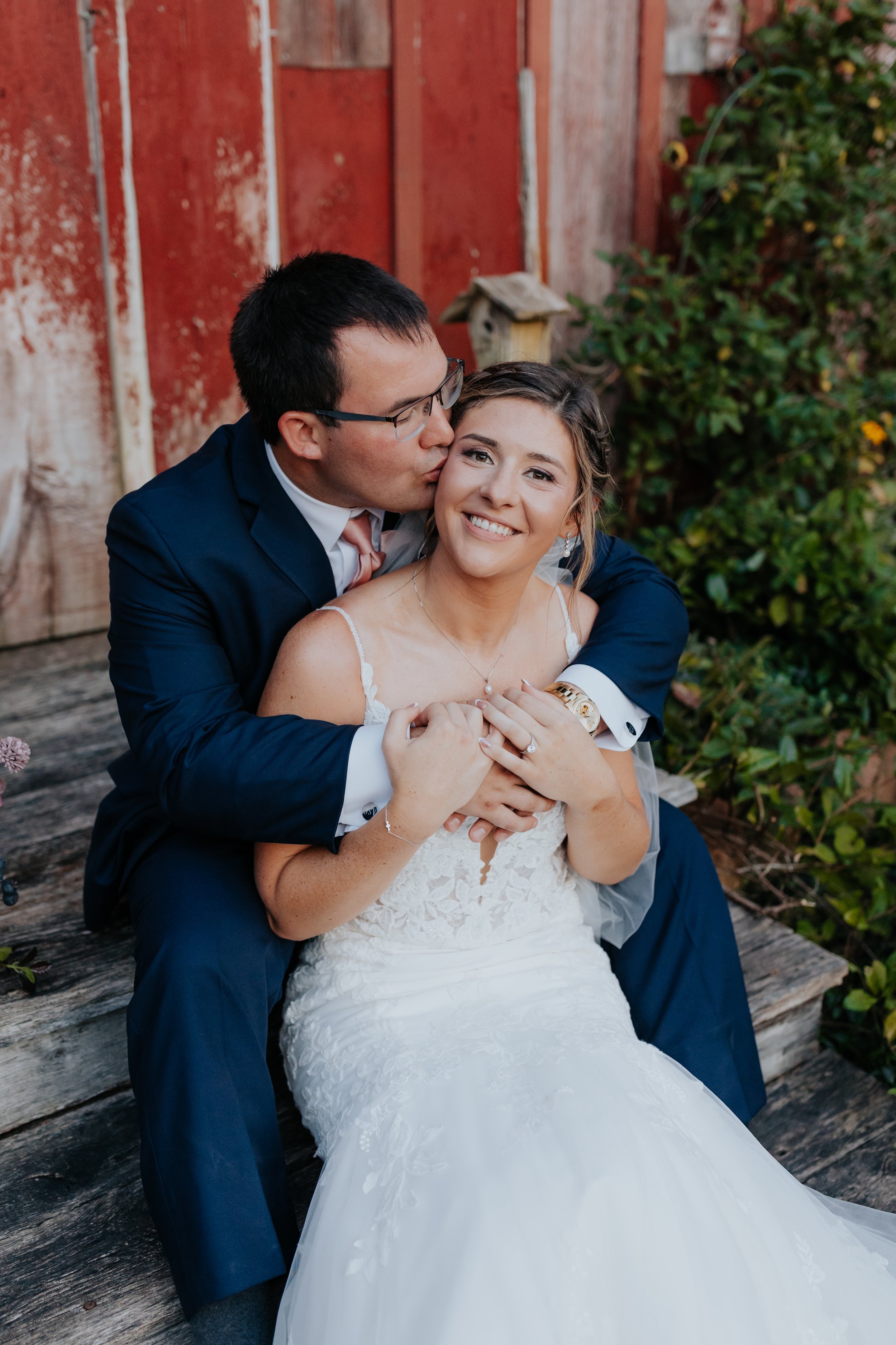 A bride and groom sitting on a wooden bench, with the groom kissing the bride on the cheek, outdoors with a red fence and green foliage in the background.