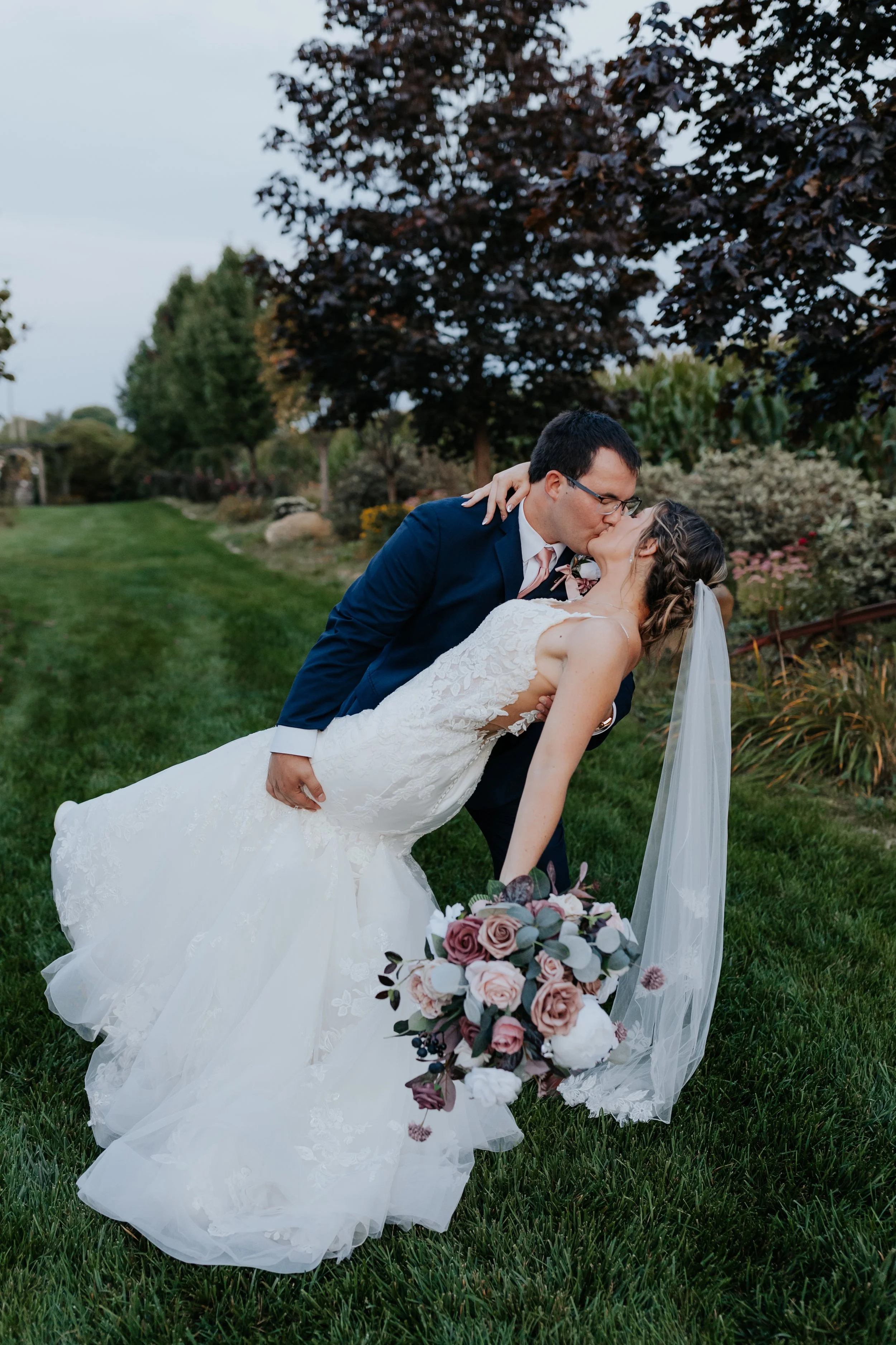 A newlywed couple sharing a kiss outdoors on a grassy field. The bride in a white wedding dress is leaning back while holding a bouquet of flowers, with her veil trailing. The groom in a dark blue suit is holding her and leaning in for a kiss. There are trees and plants in the background.
