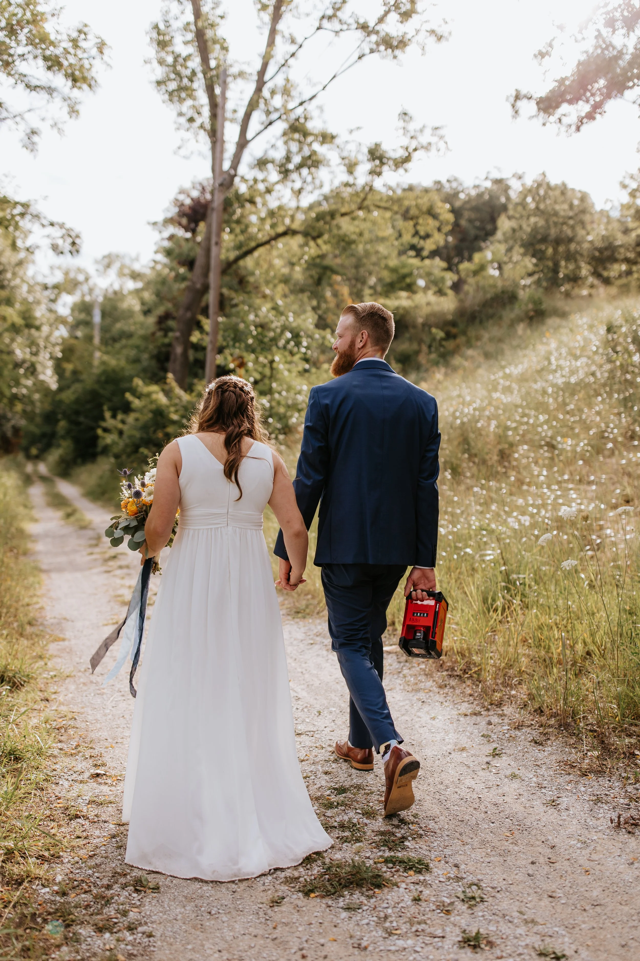A bride and groom walking hand in hand on a dirt path through nature, the bride holding a bouquet, the groom carrying a red toolbox, during sunset.