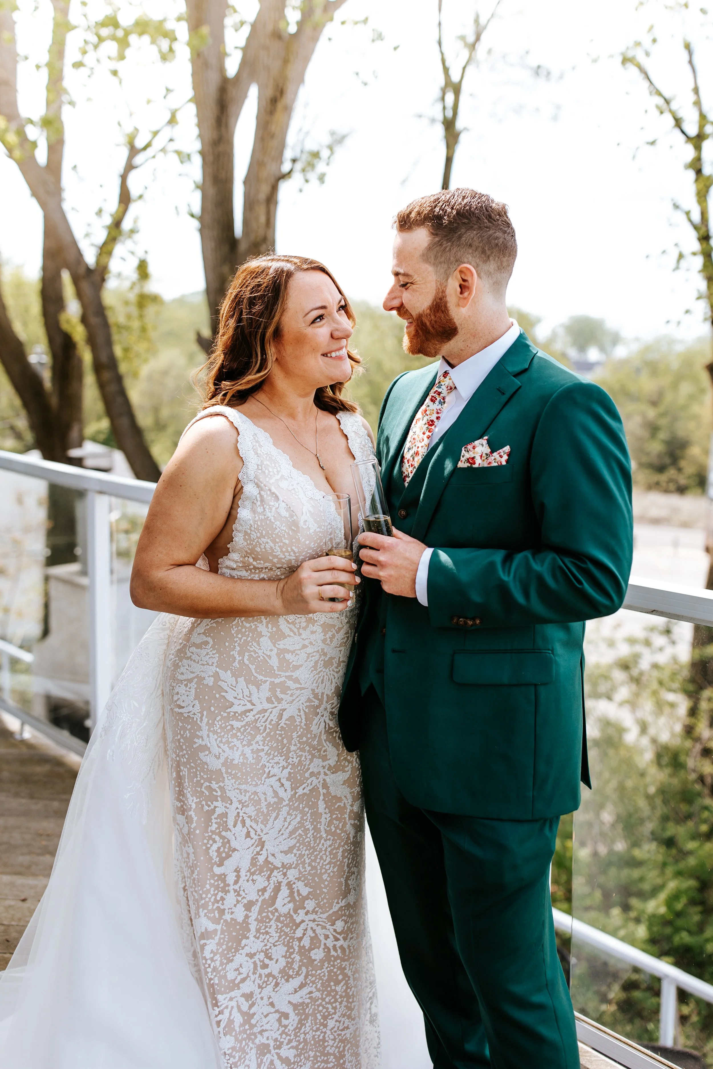 A bride and groom smiling at each other and holding champagne glasses outdoors during a wedding celebration.