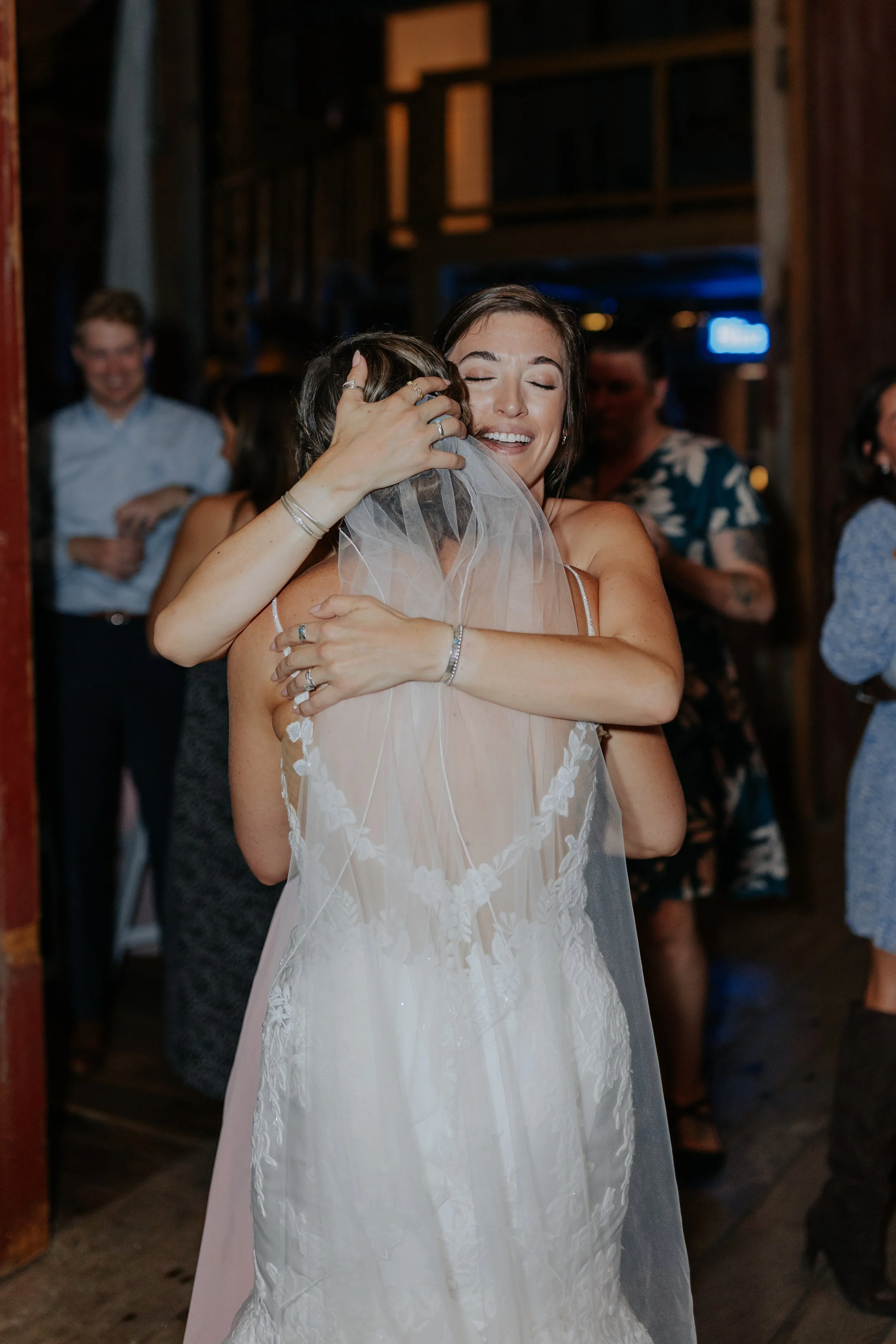 Two women hugging at a wedding celebration in a rustic venue, one in a white wedding dress with a veil, the other in a sleeveless dress, surrounded by other guests.