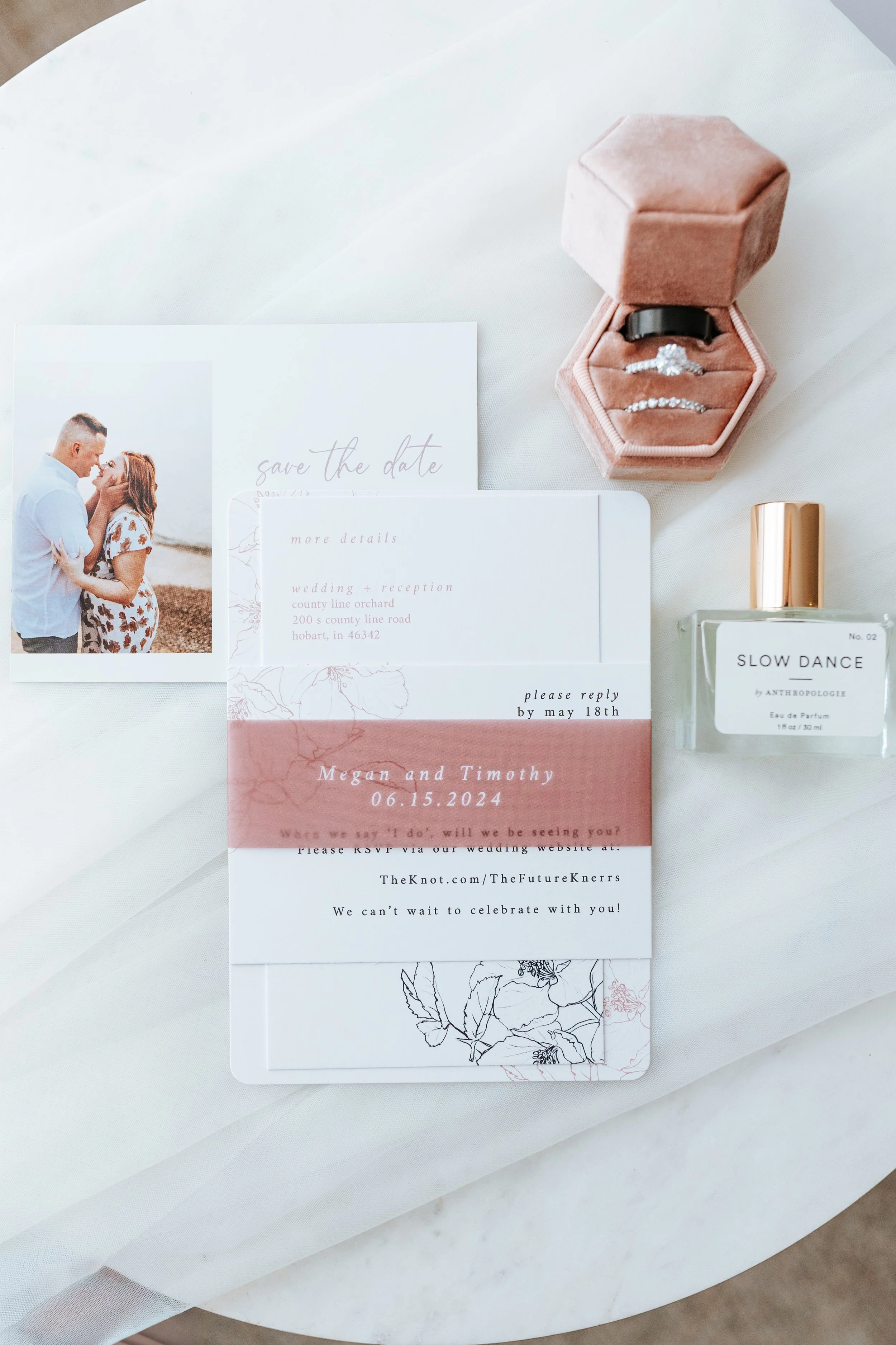 Wedding invitation suite with a photo of a couple, a save-the-date card, and RSVP details. Jewelry in a pink velvet box, perfume, and a card are also visible on a white surface.
