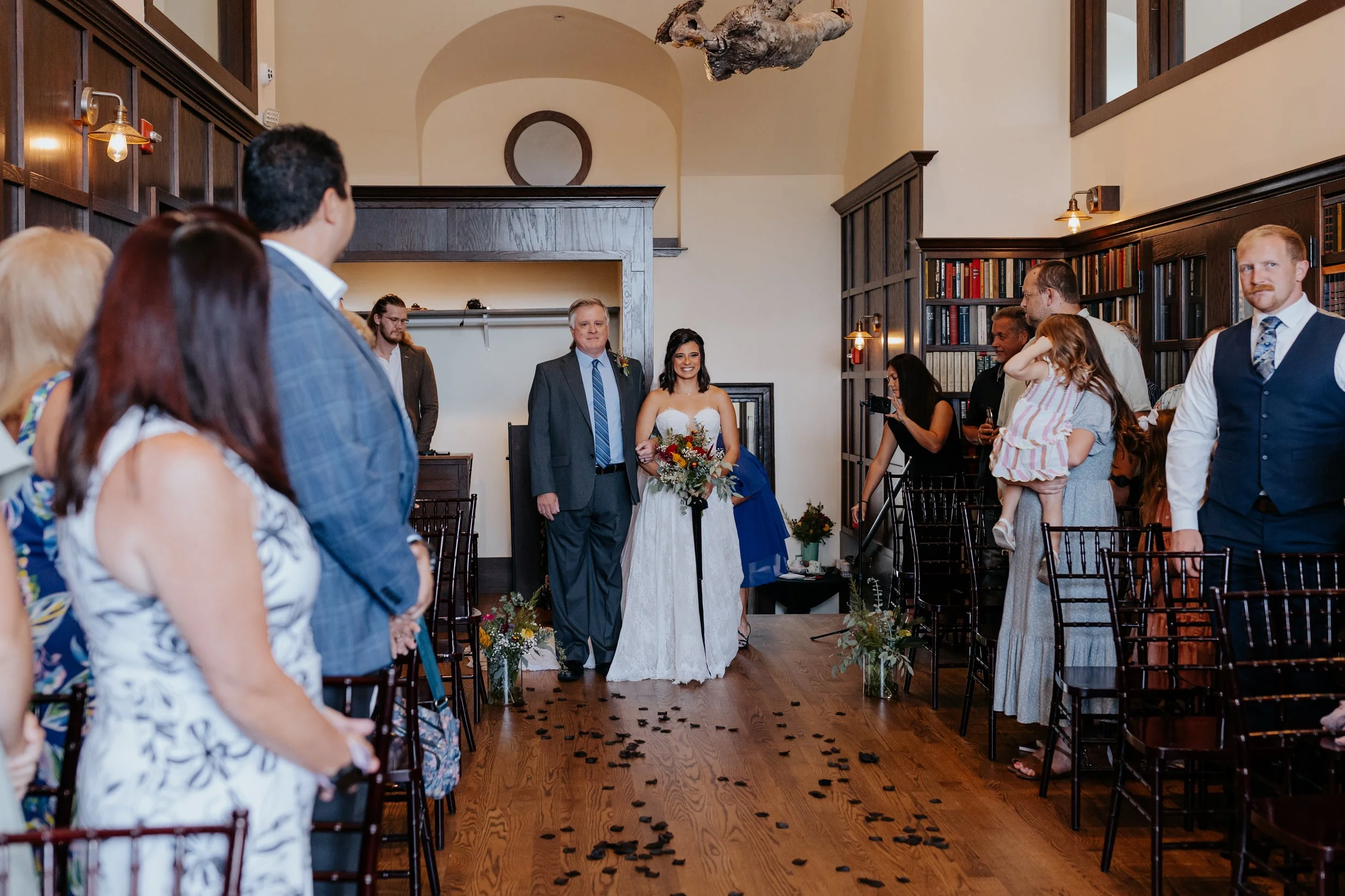 Bride walking down the aisle in a wedding ceremony, surrounded by guests in a room with wooden shelves and books, with flower arrangements on the floor.