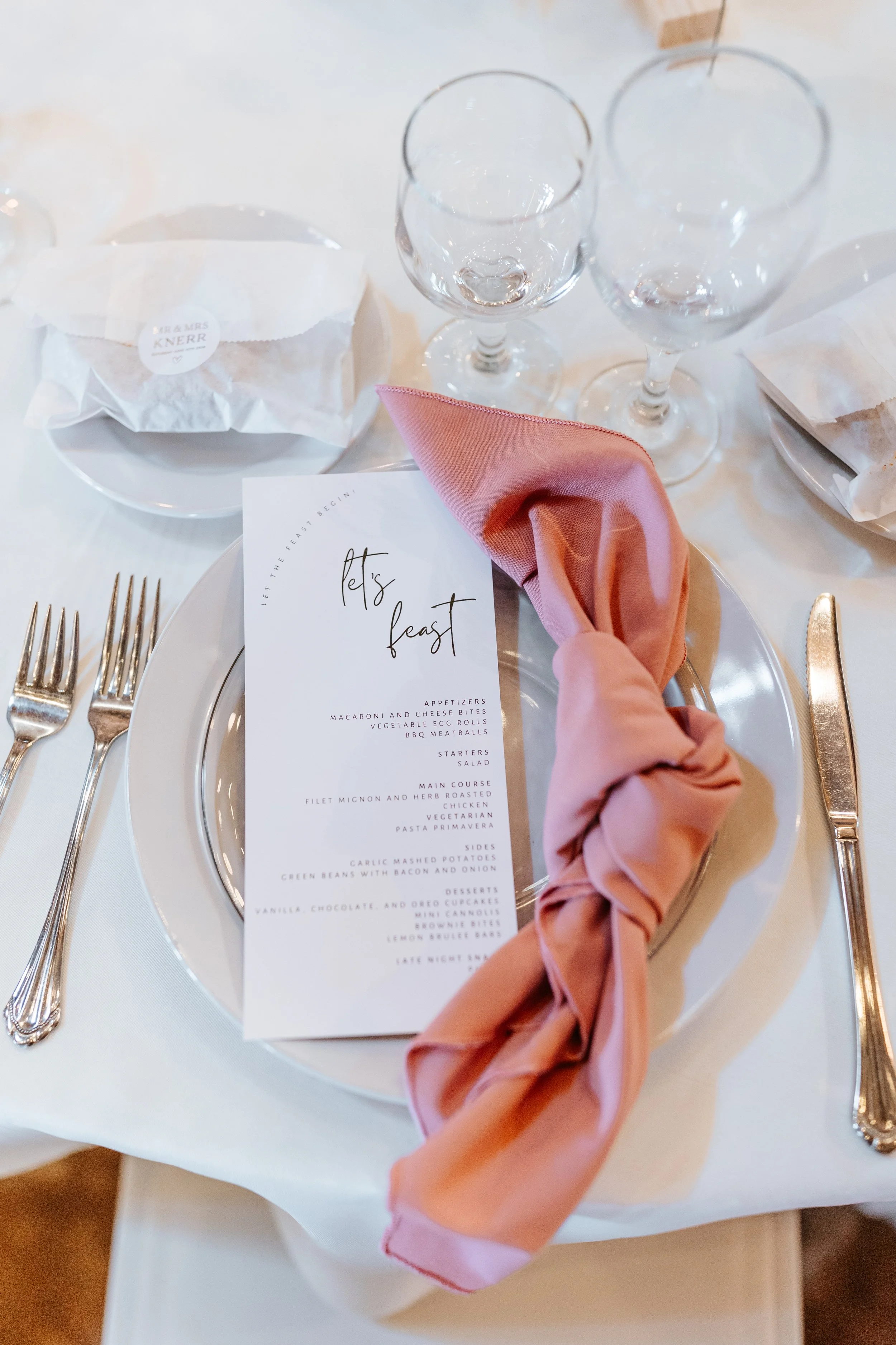 Elegant table setting with a white dinner plate, pink cloth napkin, menu, two wine glasses, and silverware, featuring a wedding or event theme.
