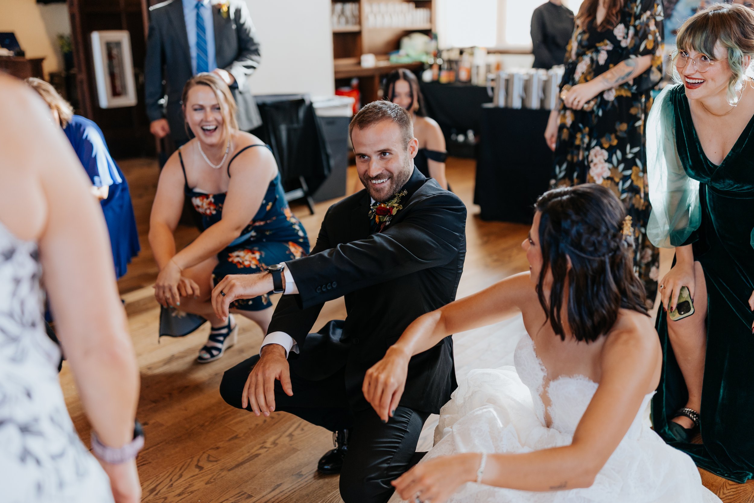 Group of wedding guests dancing and laughing indoors, including a man in a tuxedo, a bride in a white wedding dress, and women in colorful dresses.