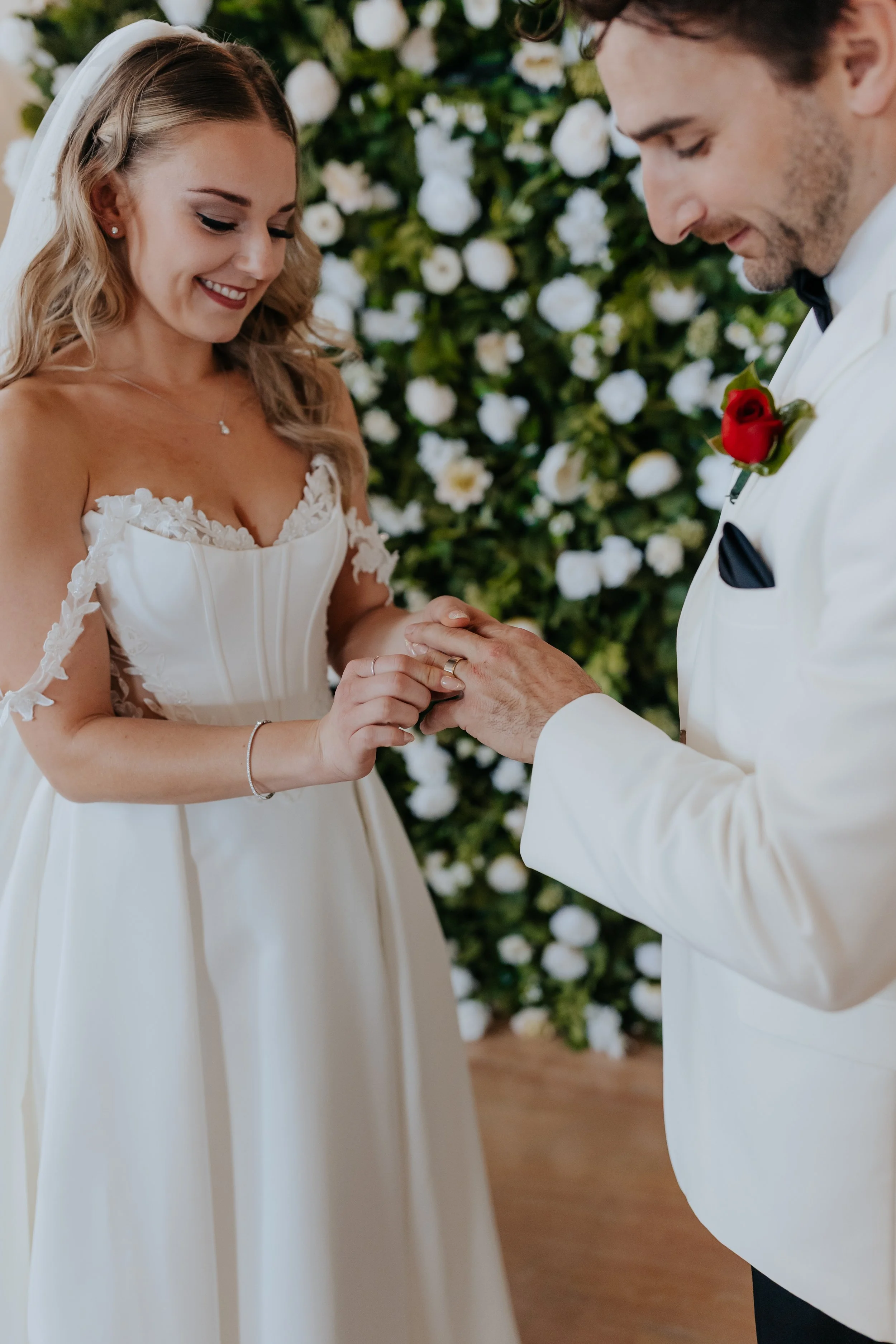 A bride and groom exchanging wedding rings during their ceremony, with a floral backdrop of white flowers.