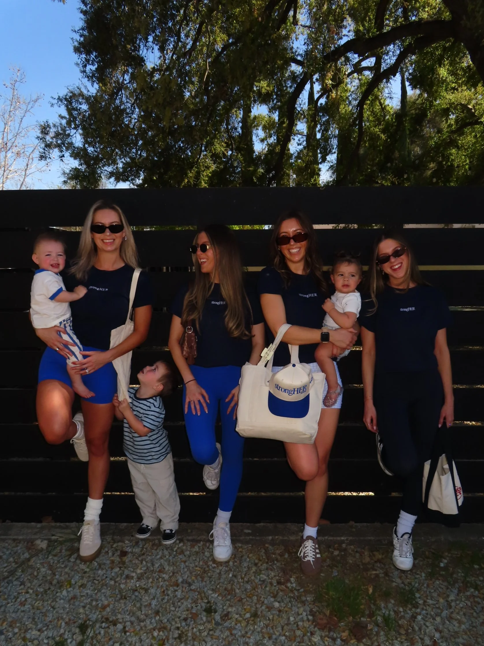 Group of five women and three children posing outdoors, wearing sunglasses and matching navy blue strongHER branded baby tees, with trees in the background.