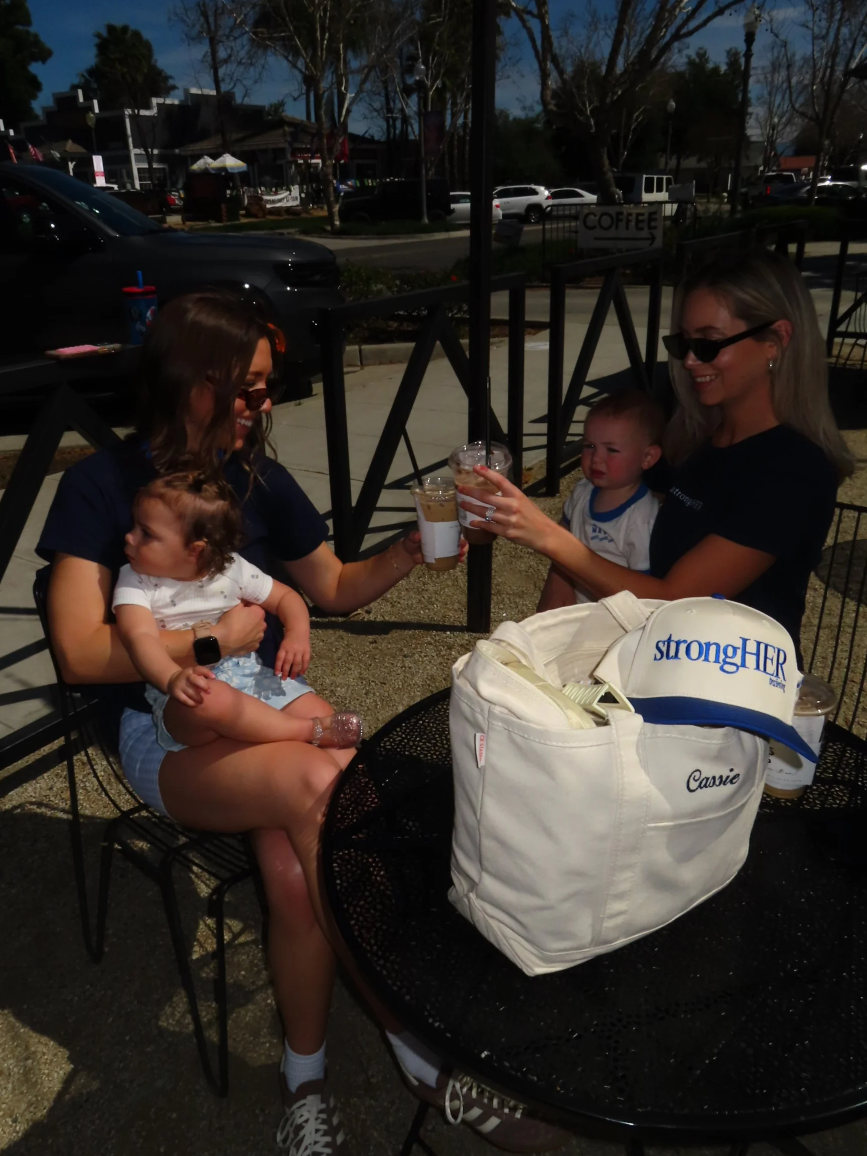 Two women with children sitting at an outdoor cafe table, clinking drinks and smiling, with a white bag and the strongHER training baseball cap on the table.