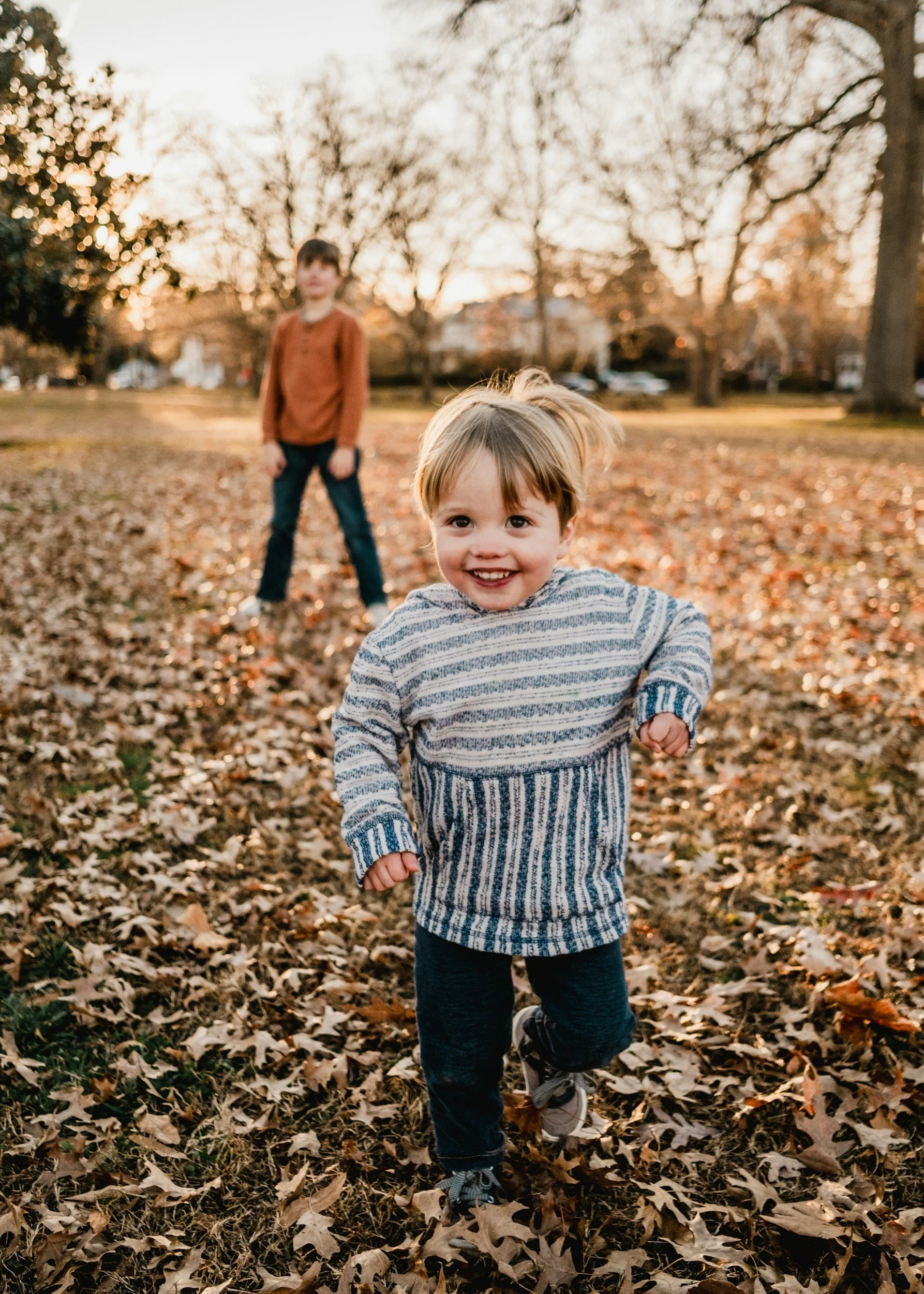 A young boy with a happy expression running through a park covered with fallen autumn leaves, with an older boy standing in the background during sunset.