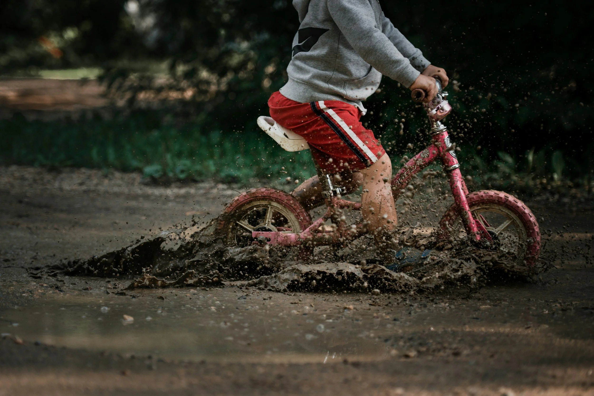 Child riding a pink bicycle through a muddy puddle.