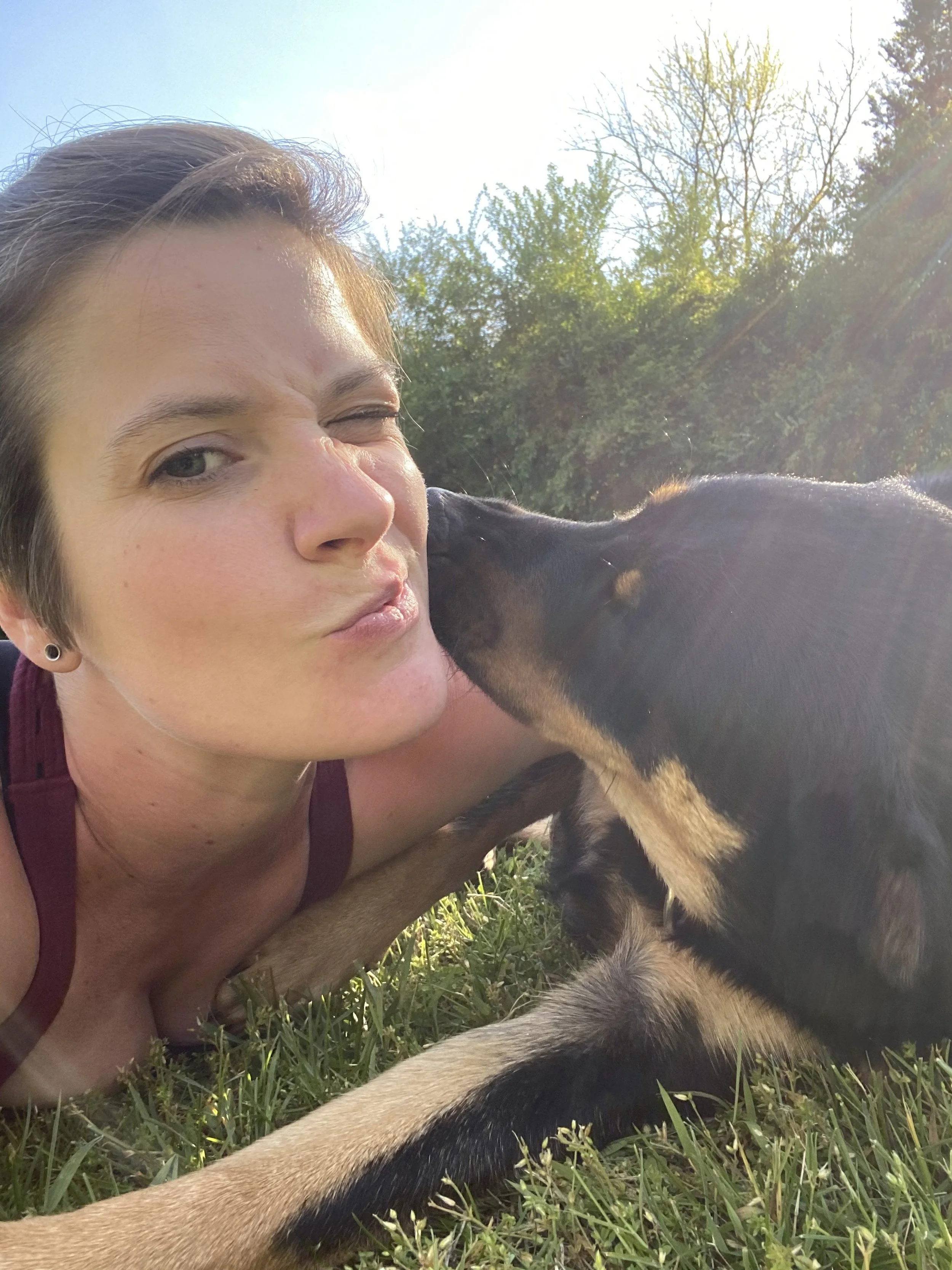 A woman lying on the grass outdoors, winking and puckering her lips while a puppy gives her a kiss.