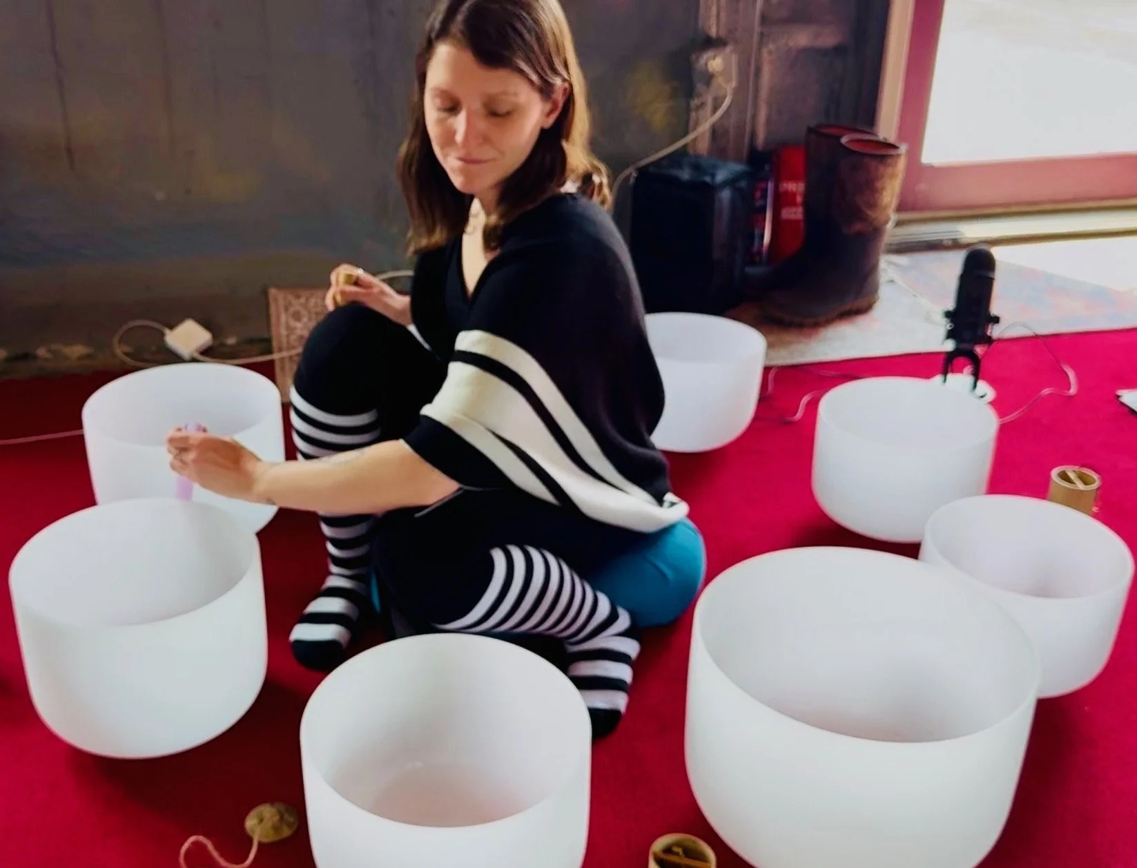 A woman sitting on a red carpet surrounded by white crystal singing bowls, preparing to play one with a mallet, in a room with wooden walls and a window.