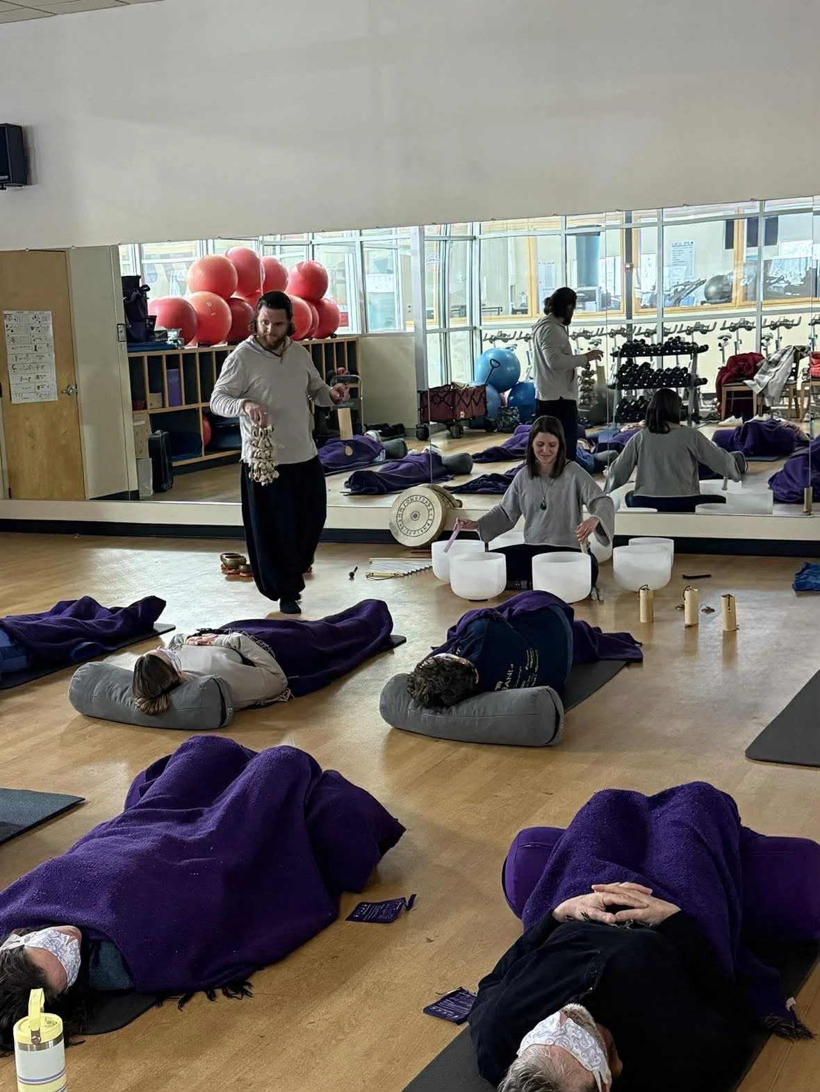 People lying on yoga mats in a group meditation or yoga class, using cushions and blankets, with some individuals wearing masks. Two instructors are playing crystal bowls and a gong, with yoga equipment and large exercise balls visible in the backgro