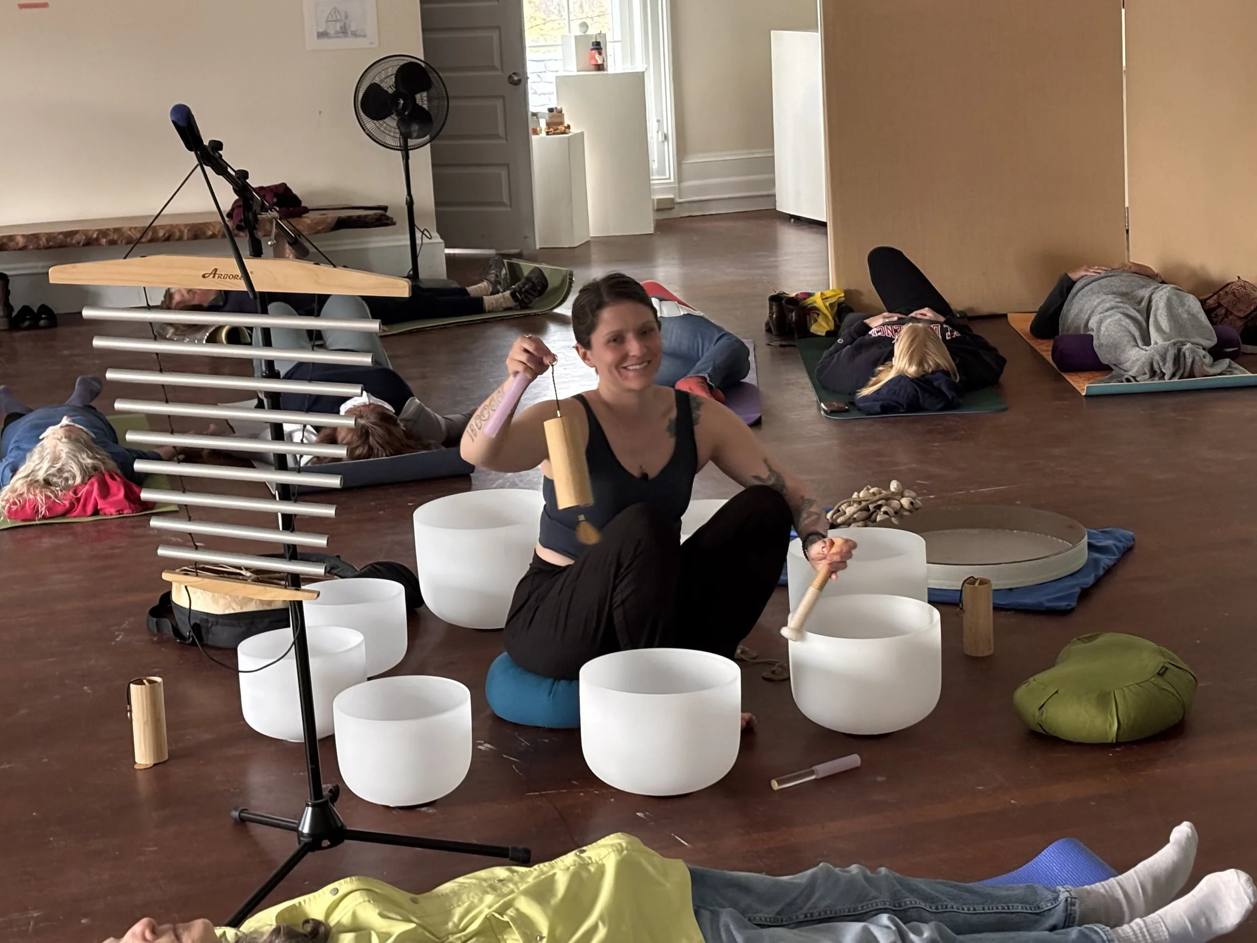 Group of people participating in a sound bath meditation session, lying on mats and sitting among singing bowls, with some holding mallets. A woman in the foreground is smiling and holding a mallet, with others resting or practicing relaxation