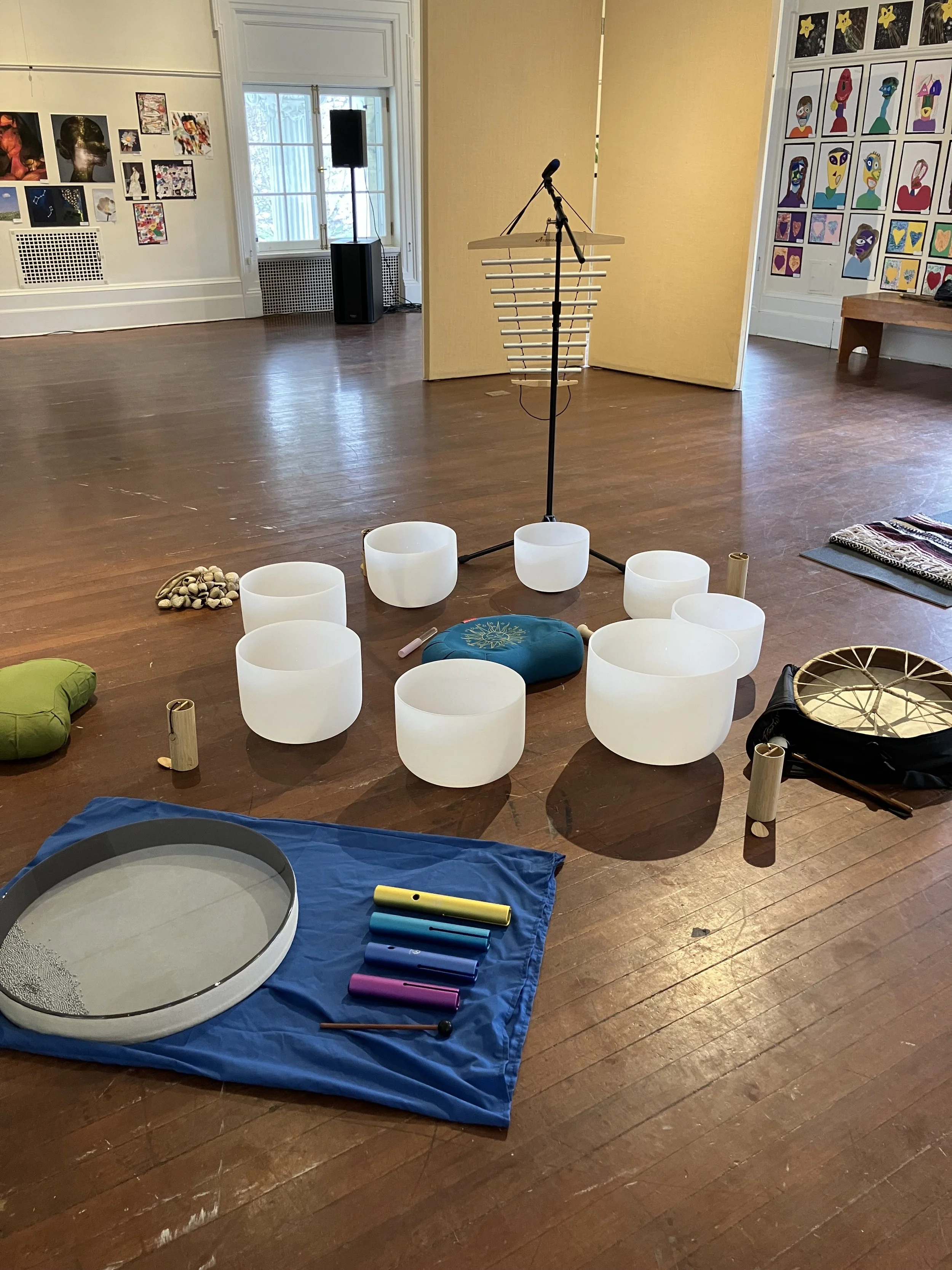 An art gallery with various artworks on the walls; a music setup with singing bowls, mallets, and a microphone stand; a green cushion, a blue cushion, and a drum on a blue mat; and colorful wind chimes on the floor.