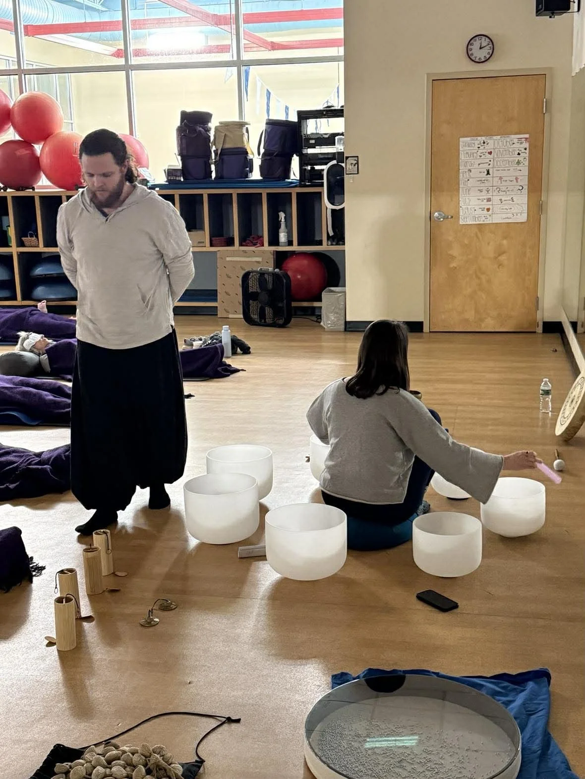 People participating in a meditation or sound healing session in a spacious room with wooden floors, large windows, and calming decor.