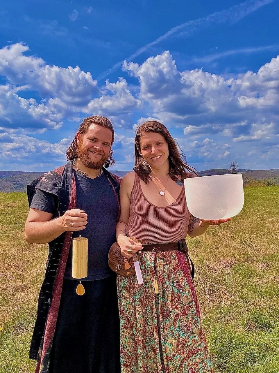 A man and woman outdoors in a grassy field under a blue sky with clouds, holding a crystal singing bowl.
