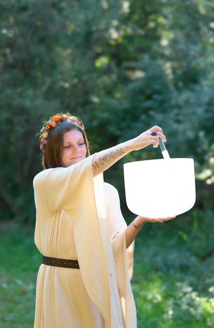 A woman with a floral crown, wearing a beige shawl and a brown belt, holding a singing bowl with a mallet in an outdoor setting with green blurred background.