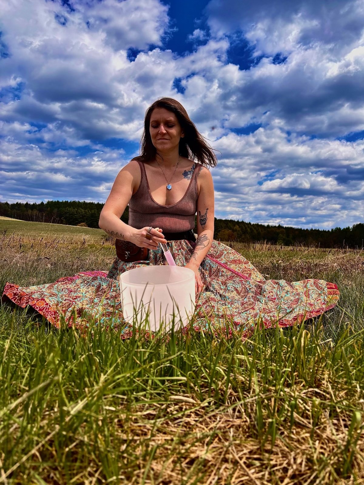 Woman sitting on a colorful blanket in a grassy field, playing a crystal singing bowl with a mallet, under a partly cloudy sky.