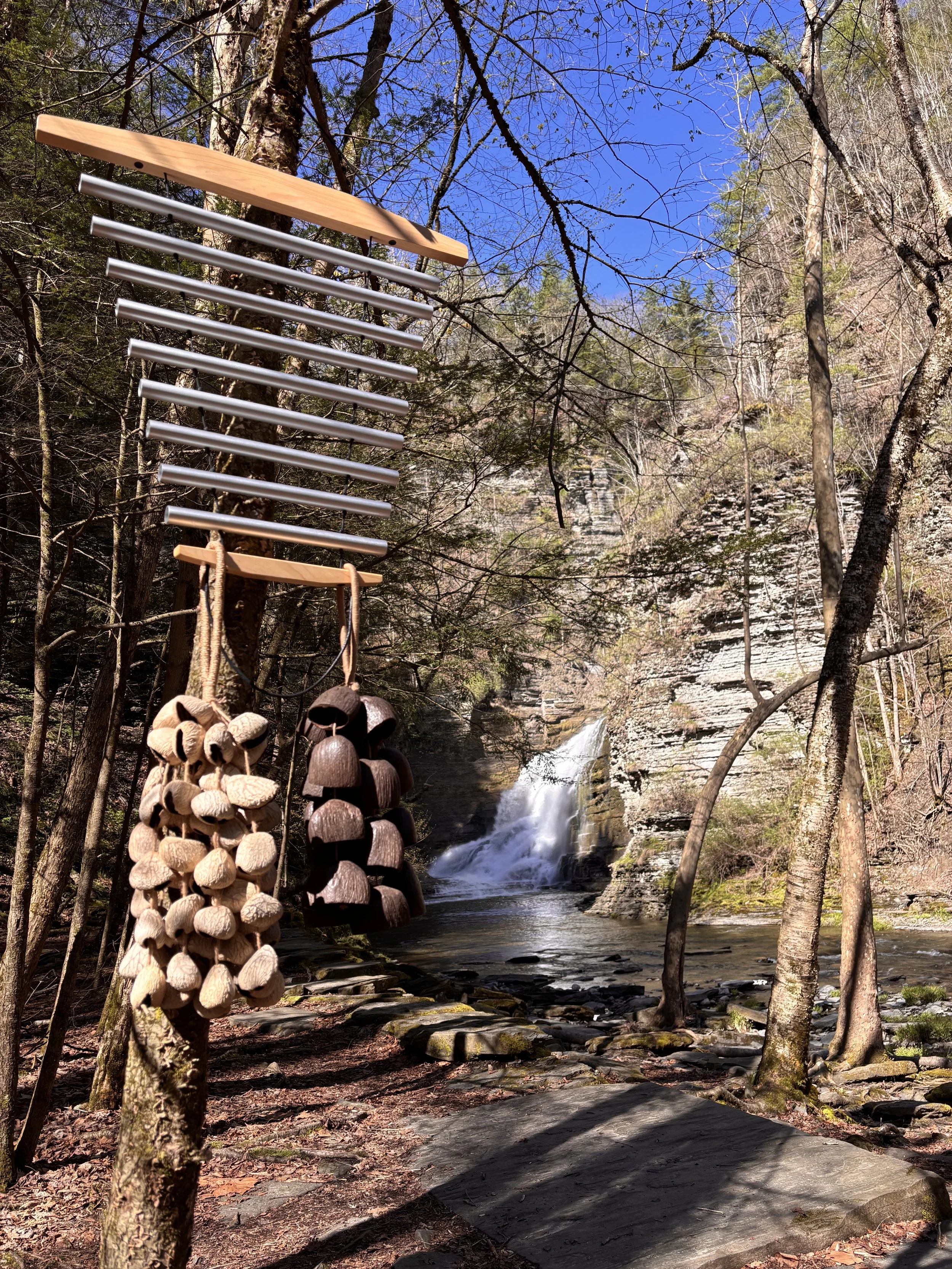A forest scene with a waterfall flowing over rocks, surrounded by trees. In the foreground, there are decorative wind chimes hanging from a tree branch, made of wood and metal.