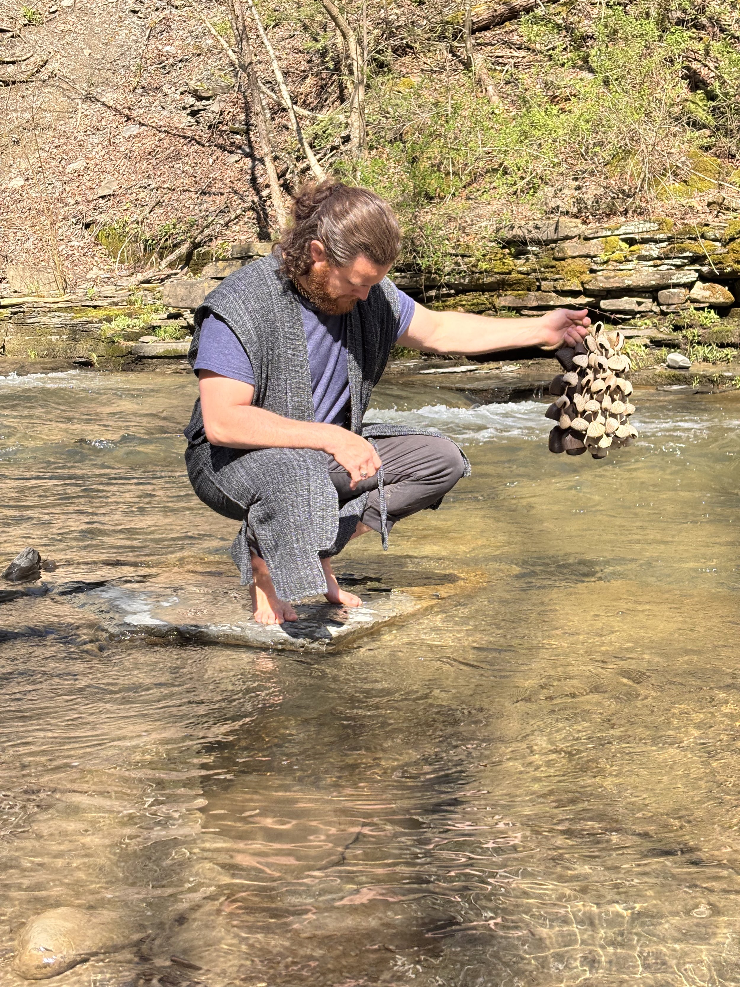 A man with long hair and a beard sits in a shallow creek, holding a rattle in his right hand, with trees and rocks in the background.