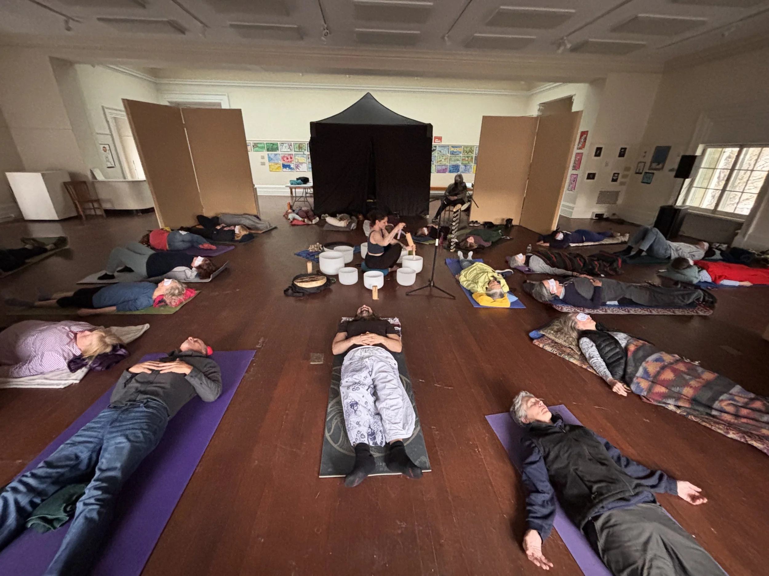 A group of people lying on yoga mats in a room, participating in a relaxing or meditative activity with a black tent-like structure in the background.