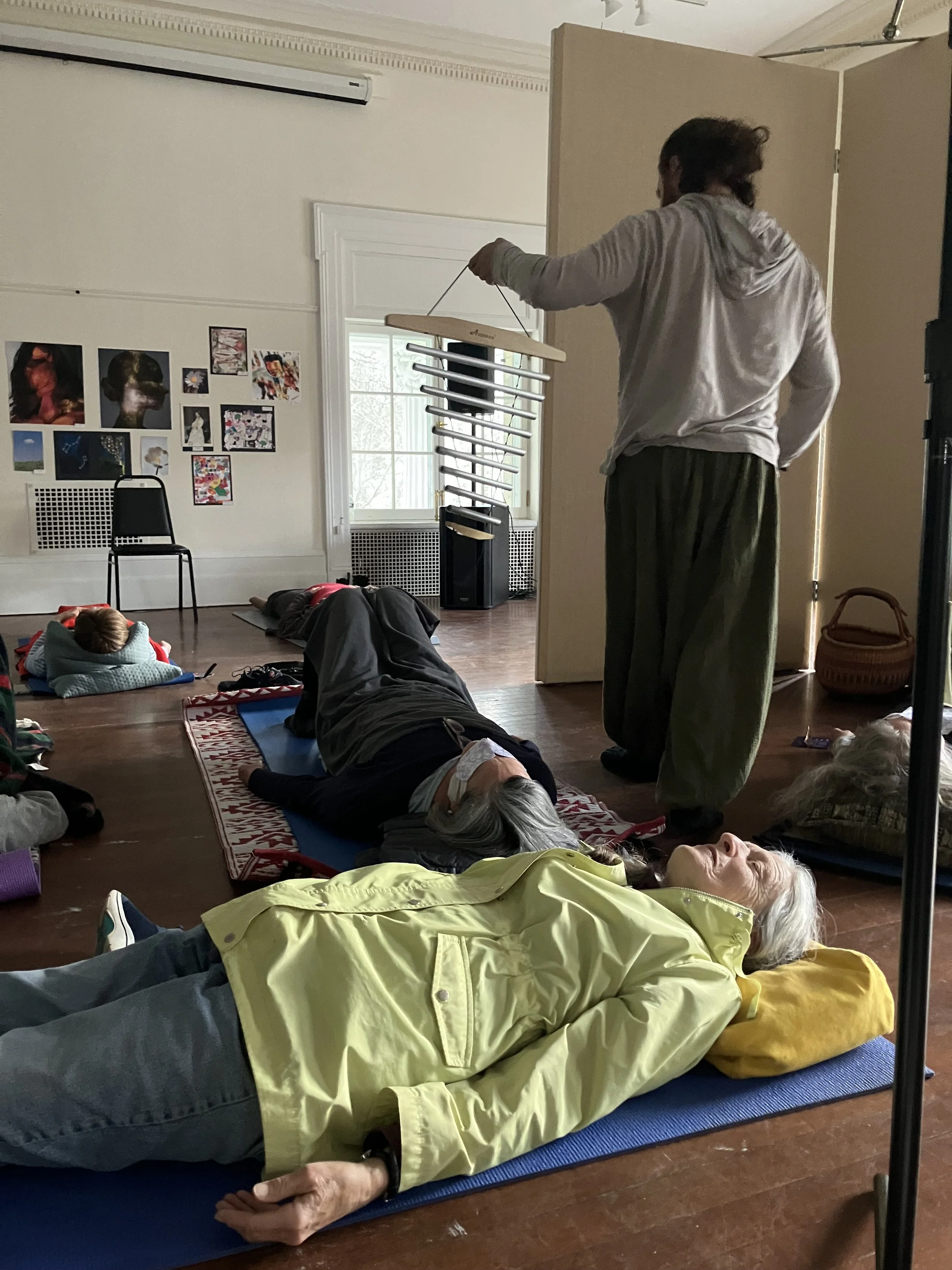 People lying on yoga mats practicing relaxation or meditation inside a room with white walls, artworks on the wall, and a window.