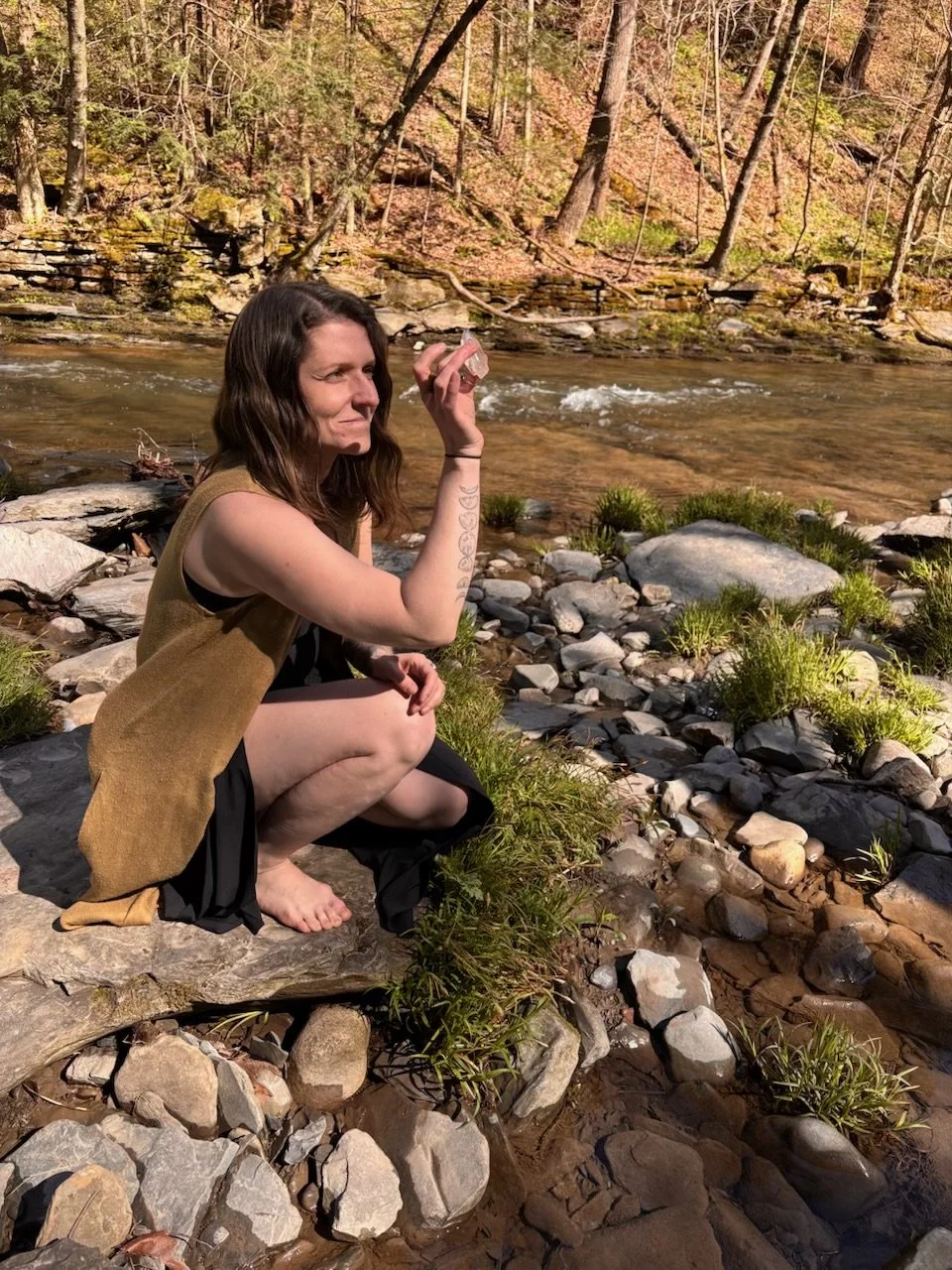 A woman with long brown hair squatting on rocks by a stream in a forest, holding a crystal in her hand.