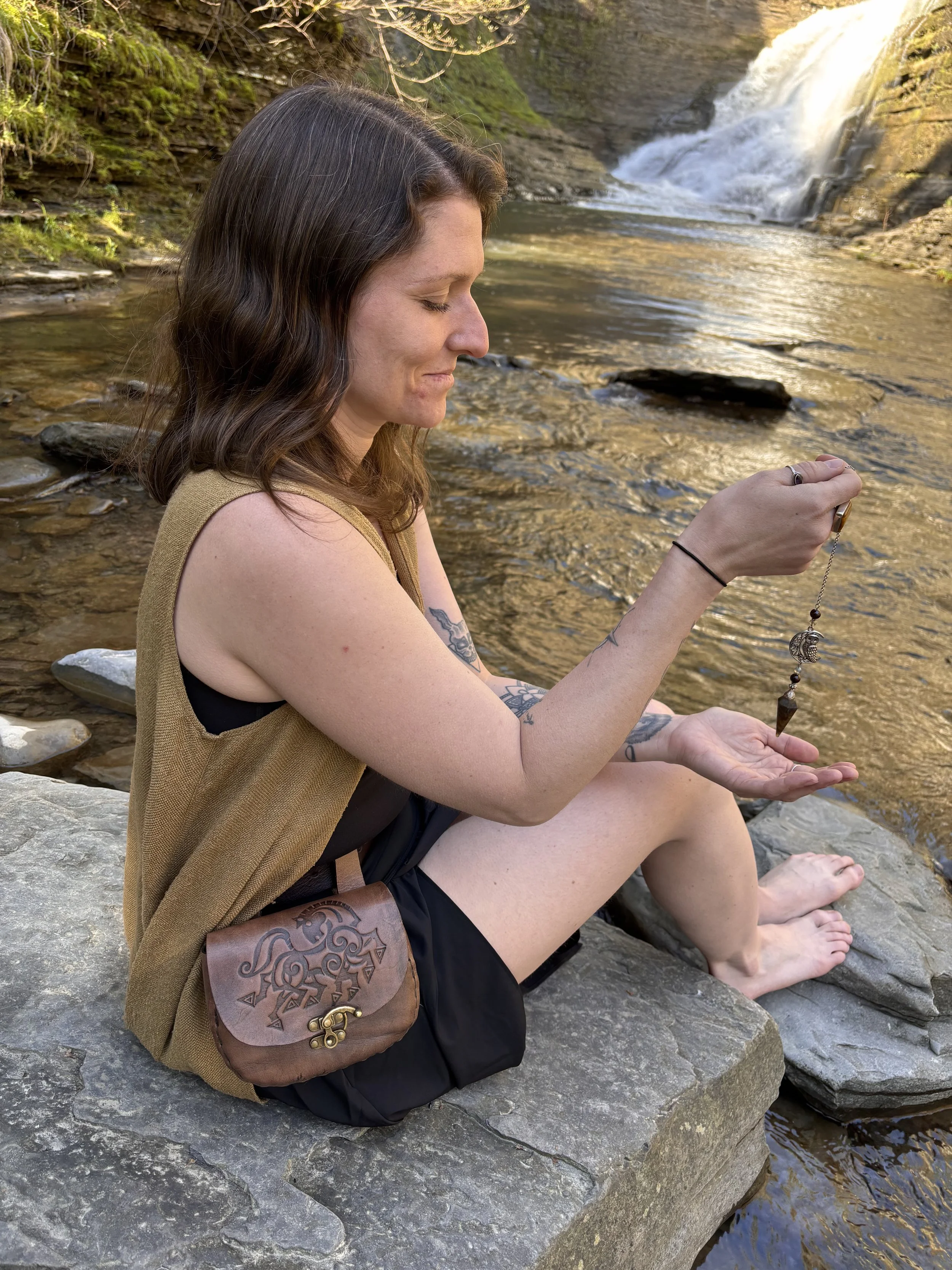A woman with long dark hair sitting on rocks by a river with a waterfall in the background, holding a necklace with a pendant.