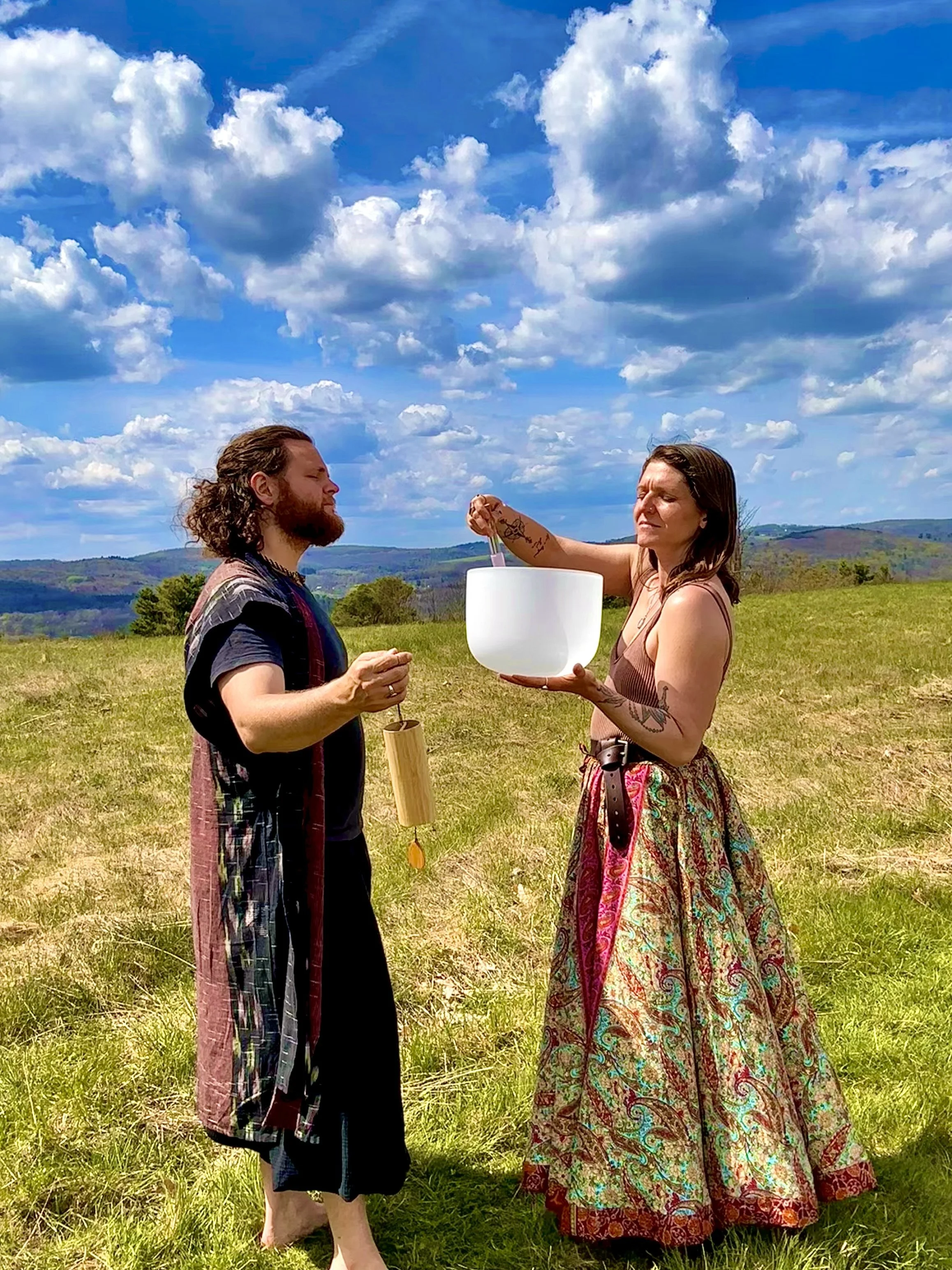 A woman and a man standing in a grassy field, the woman holding a large white singing bowl, and the man holding a mallet, with a scenic background of rolling hills and a partly cloudy sky.