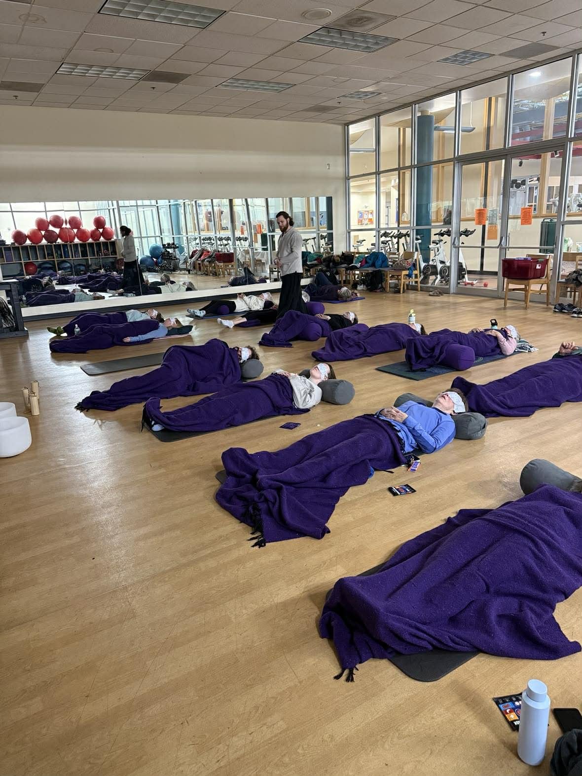 People lying on yoga mats in a fitness studio during a class, all wearing purple blankets and face masks, with a large mirror and gym equipment visible in the background.