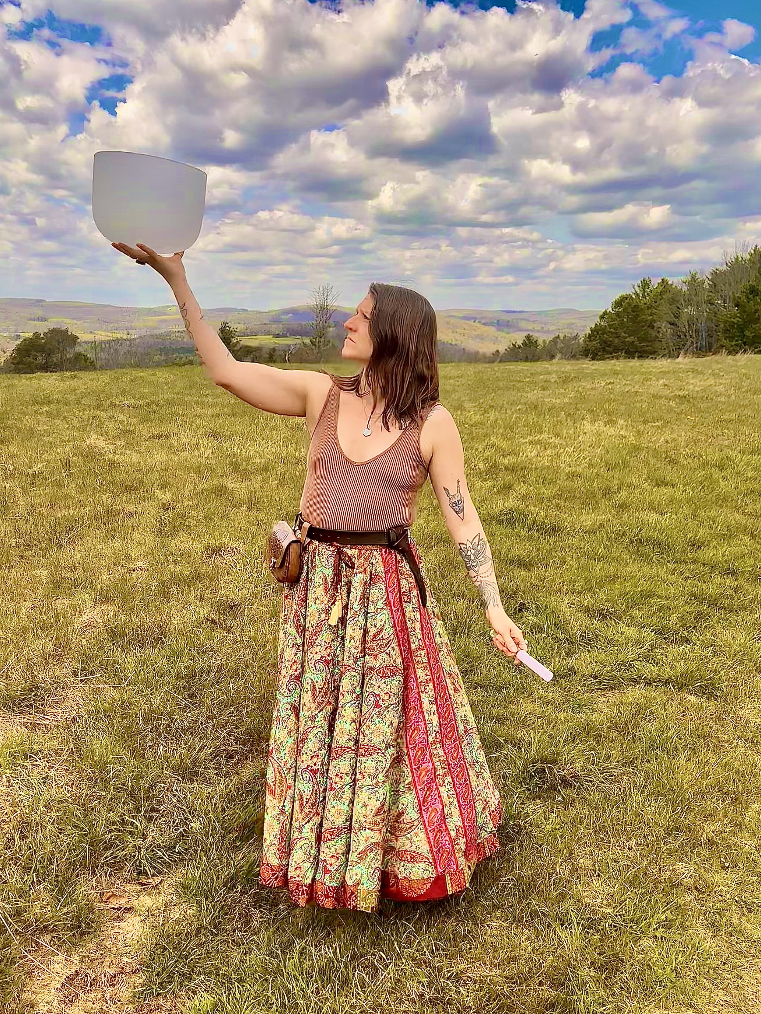 A woman standing outdoors on a grassy field, holding a large white bowl or container high in the air with her right hand, with clouds and hills in the background.