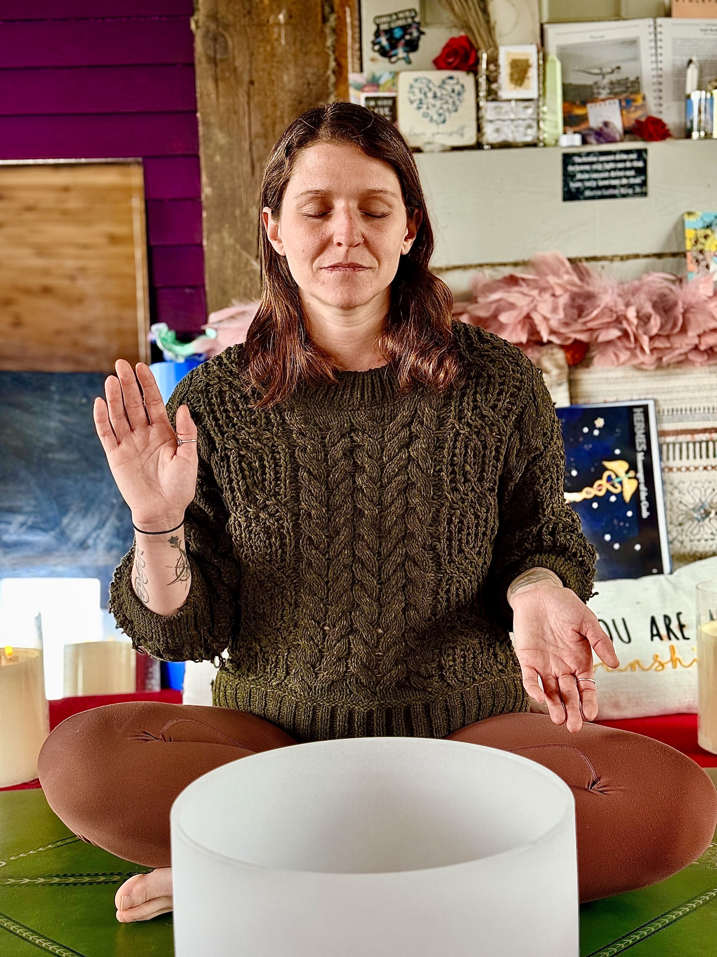 A woman with brown hair, closed eyes, and a peaceful expression is raising her right hand with palm facing forward. She is seated at a table with a large decorative platter in front of her, and her left hand is resting on the table. The background in
