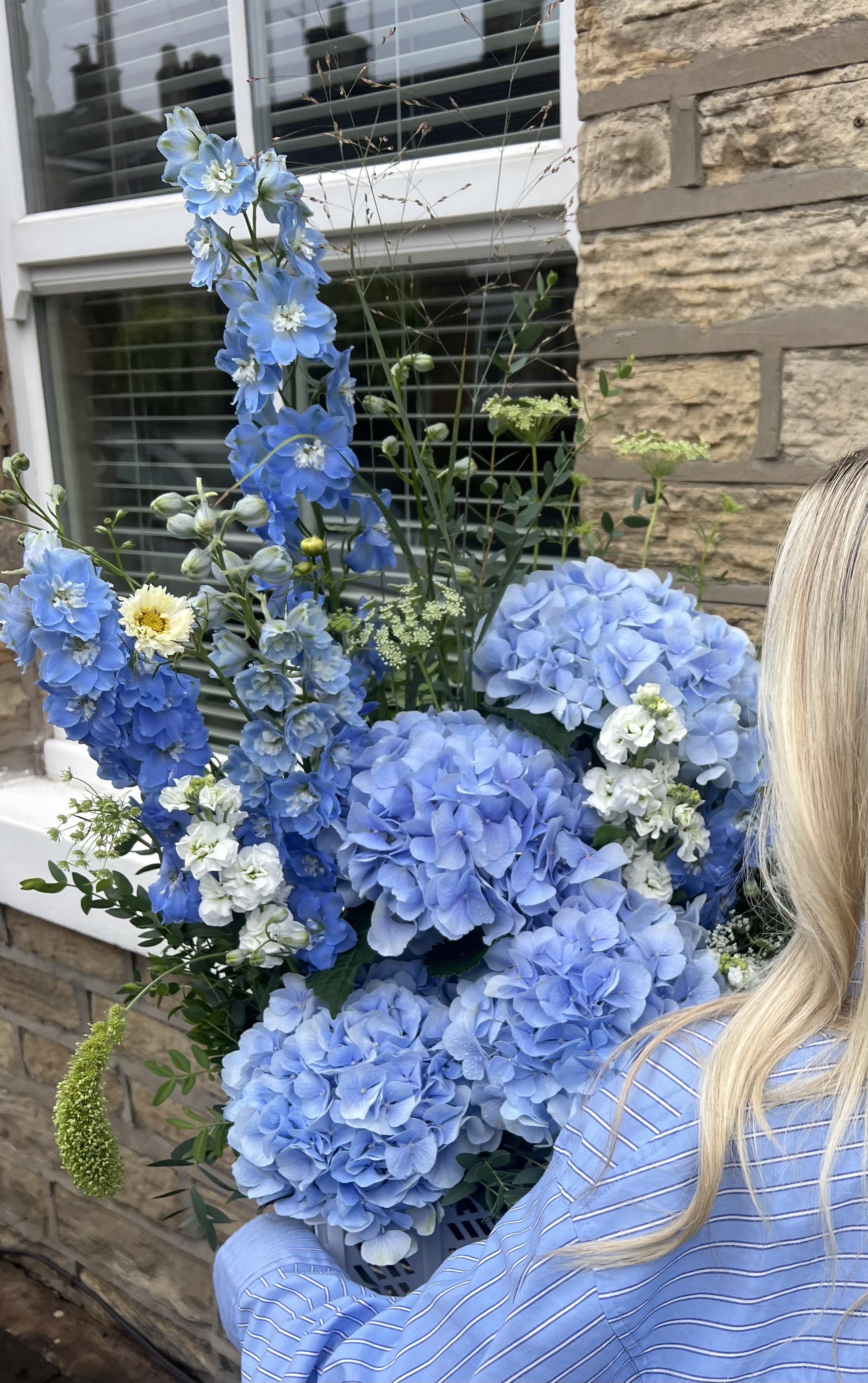 A woman holding a large bouquet of blue and white hydrangeas and delphiniums outside near a brick house with a window.