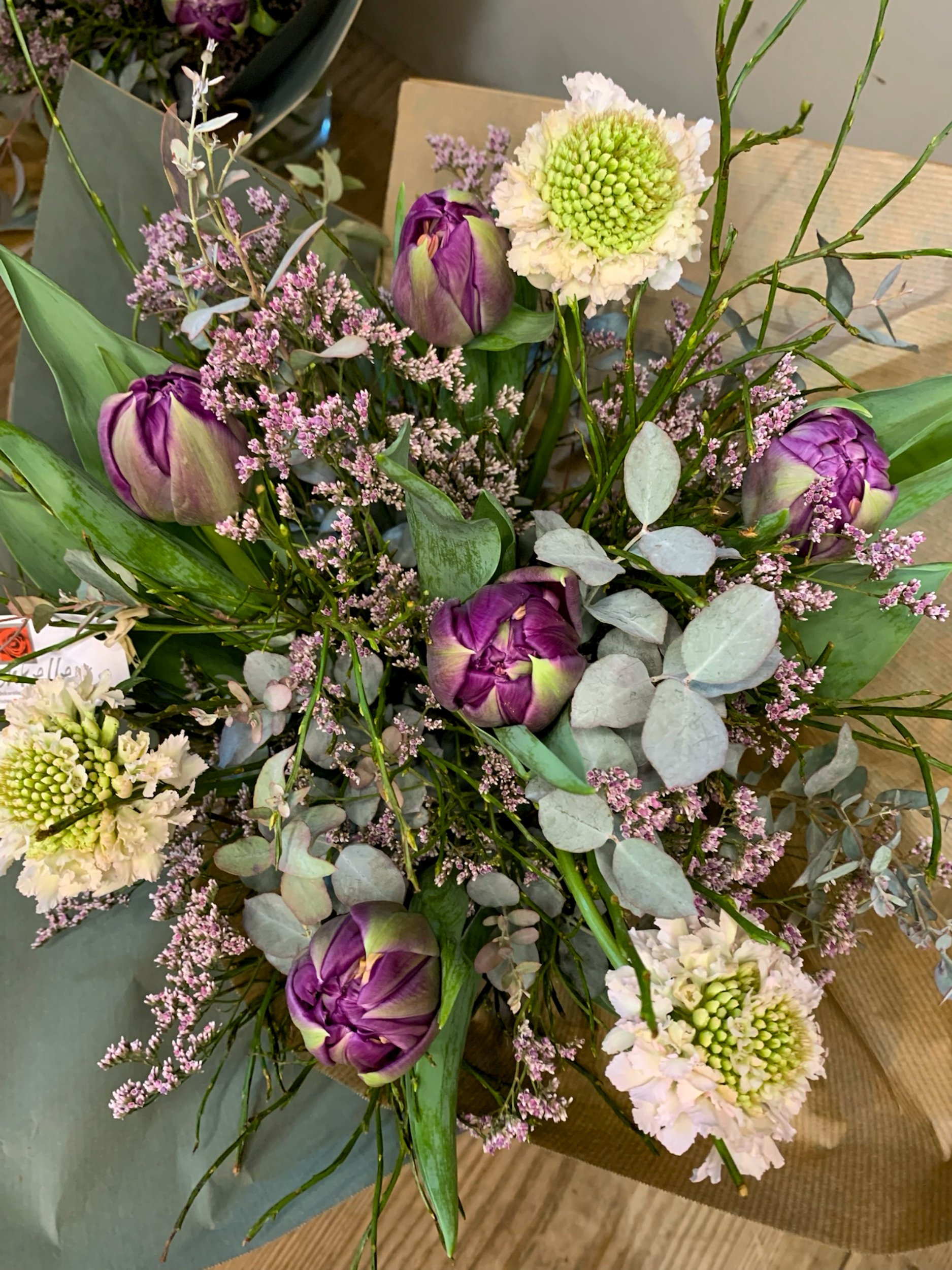 Blumenstrauß mit weißen, pinken und lila Blumen, umgeben von grünen Blättern und Zweigen, auf einem Holztisch.